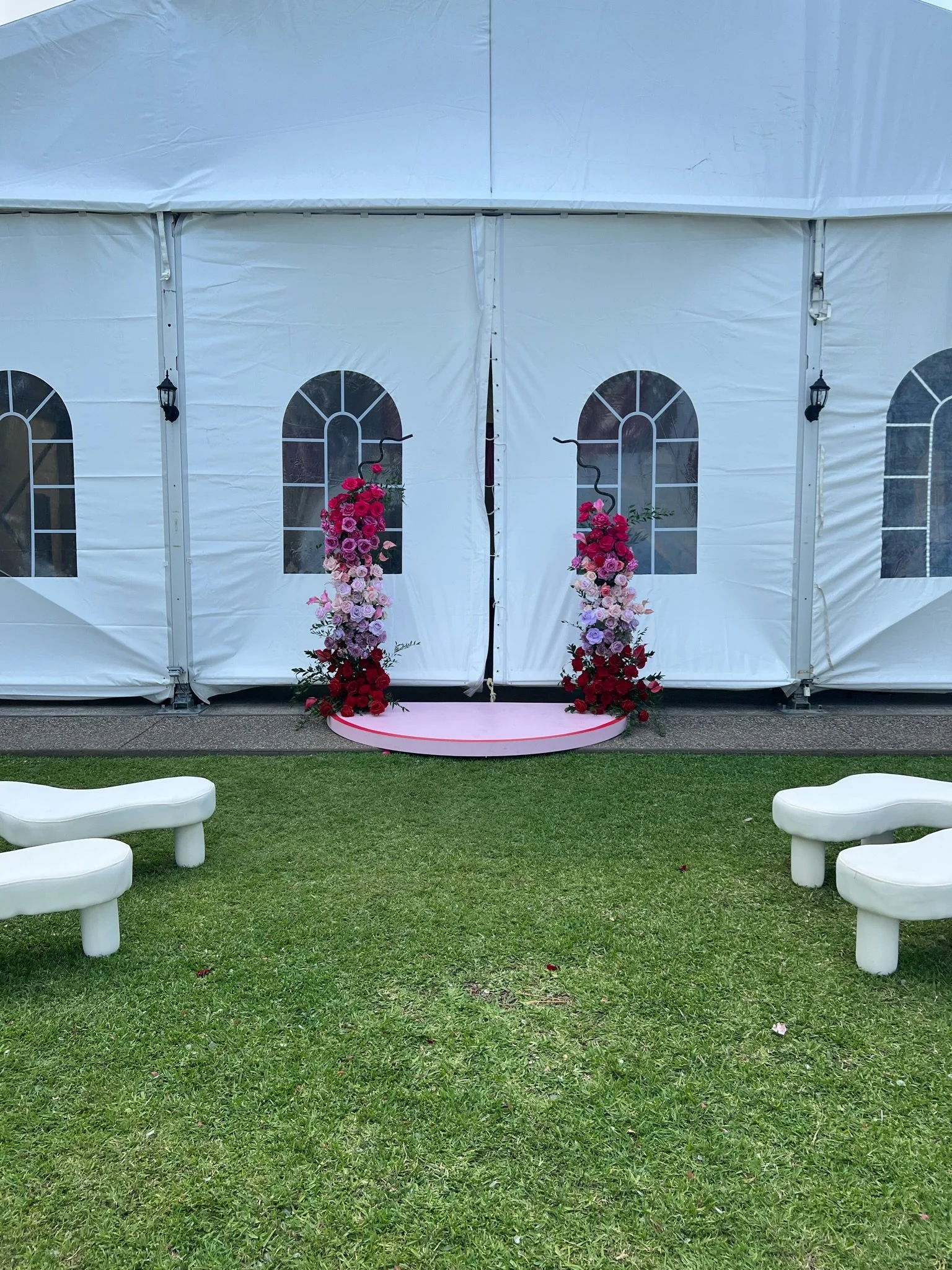 A white tent with arched windows and a small pink stage decorated with pink and red flowers for a wedding or event.