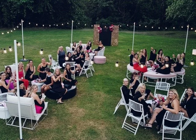Outdoor party scene with women in formal attire sitting at tables and on lounge chairs, decorated with string lights and flowers, on a grassy area.
