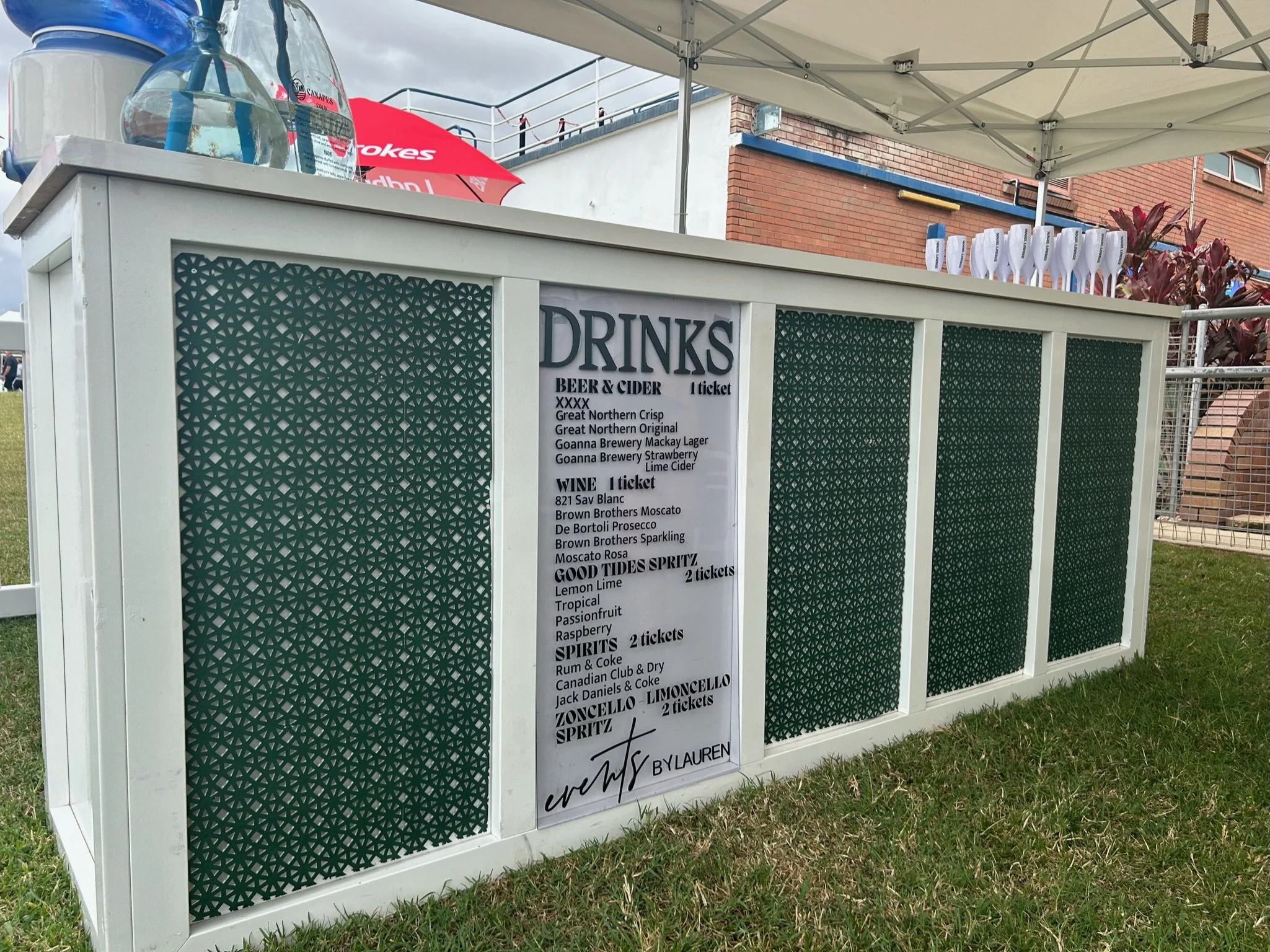 Outdoor drink stand with a menu listing beers, ciders, wines, and spirits. The stand has a decorative green pattern on its sides and is set on grass, under a white canopy.
