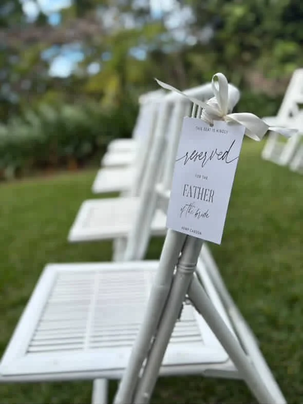 White folding chairs set up outdoors on a grassy lawn with a sign that reads 'reserved for the father of the bride' attached to a chair.