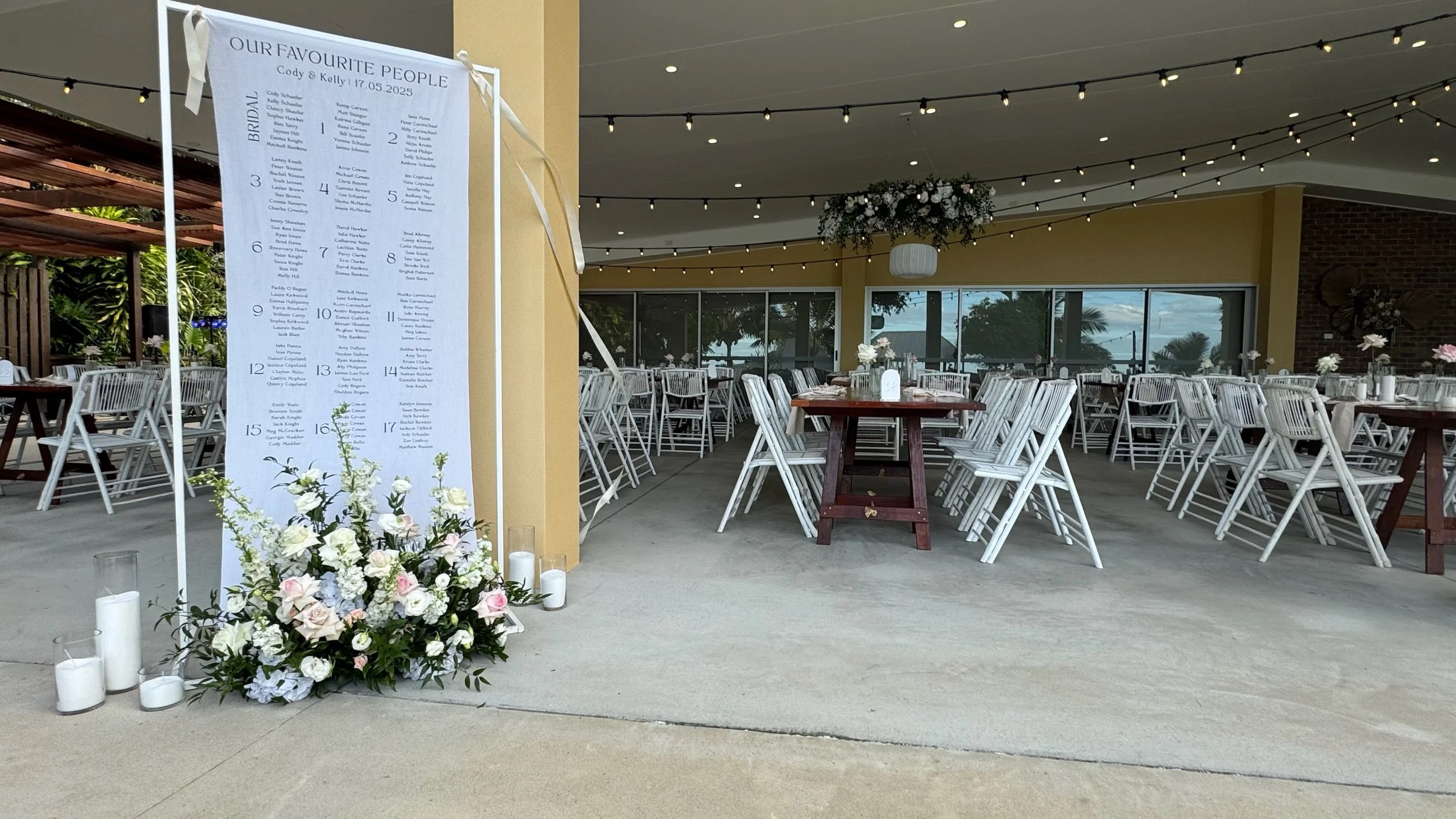 Wedding reception setup with round tables and white chairs, floral centerpieces, a large seating chart display with flowers and candles at the base, and string lights hanging from the ceiling.
