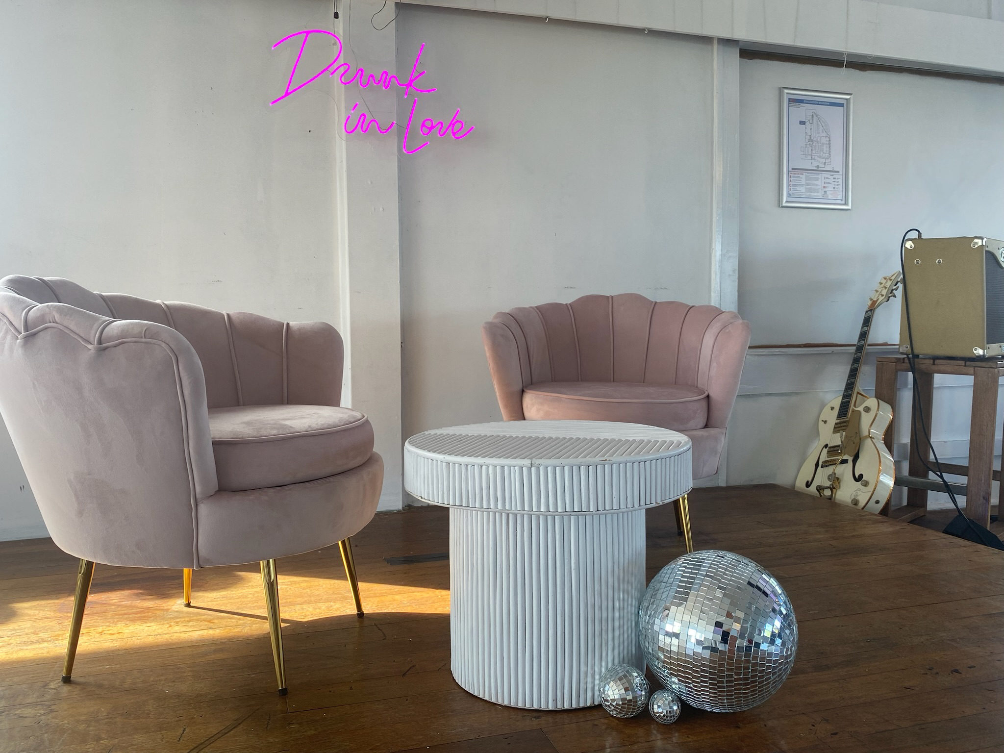A cozy lounge area with two pink velvet armchairs with gold legs, a white textured round side table, and three disco balls of different sizes on the floor. In the background, a neon sign in pink says 'Drunk in Love,' along with a guitar, an amplifier