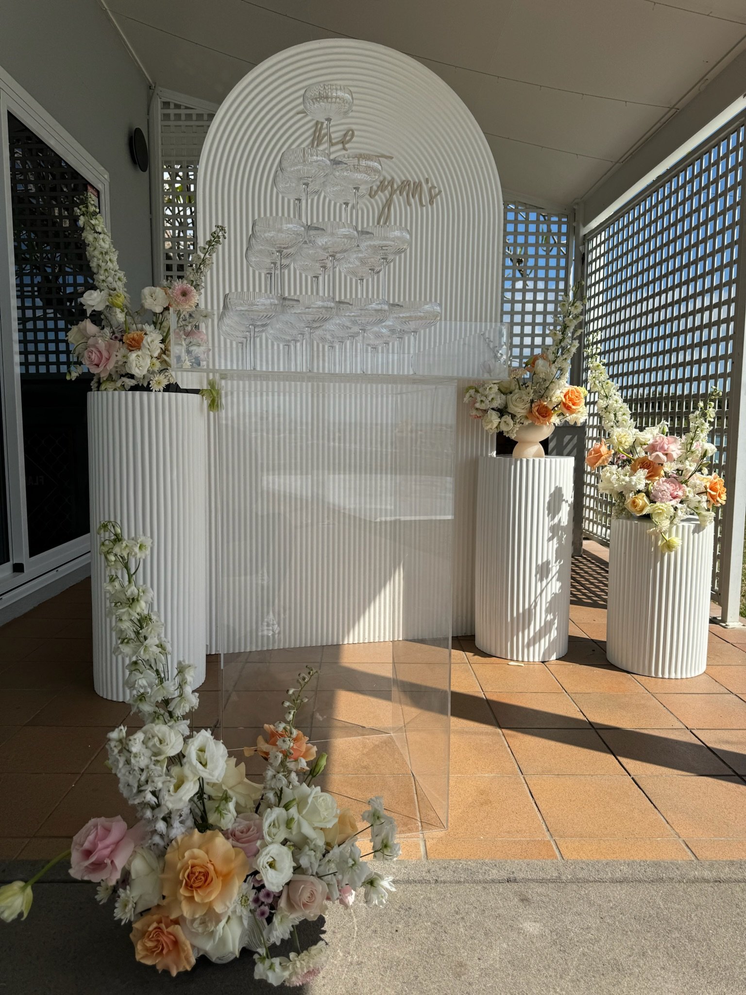 Wedding floral display with champagne glasses arranged in a pyramid, white and peach flowers in vases, a clear acrylic sign holder in front, and a backdrop with the word 'Marry' in gold script.