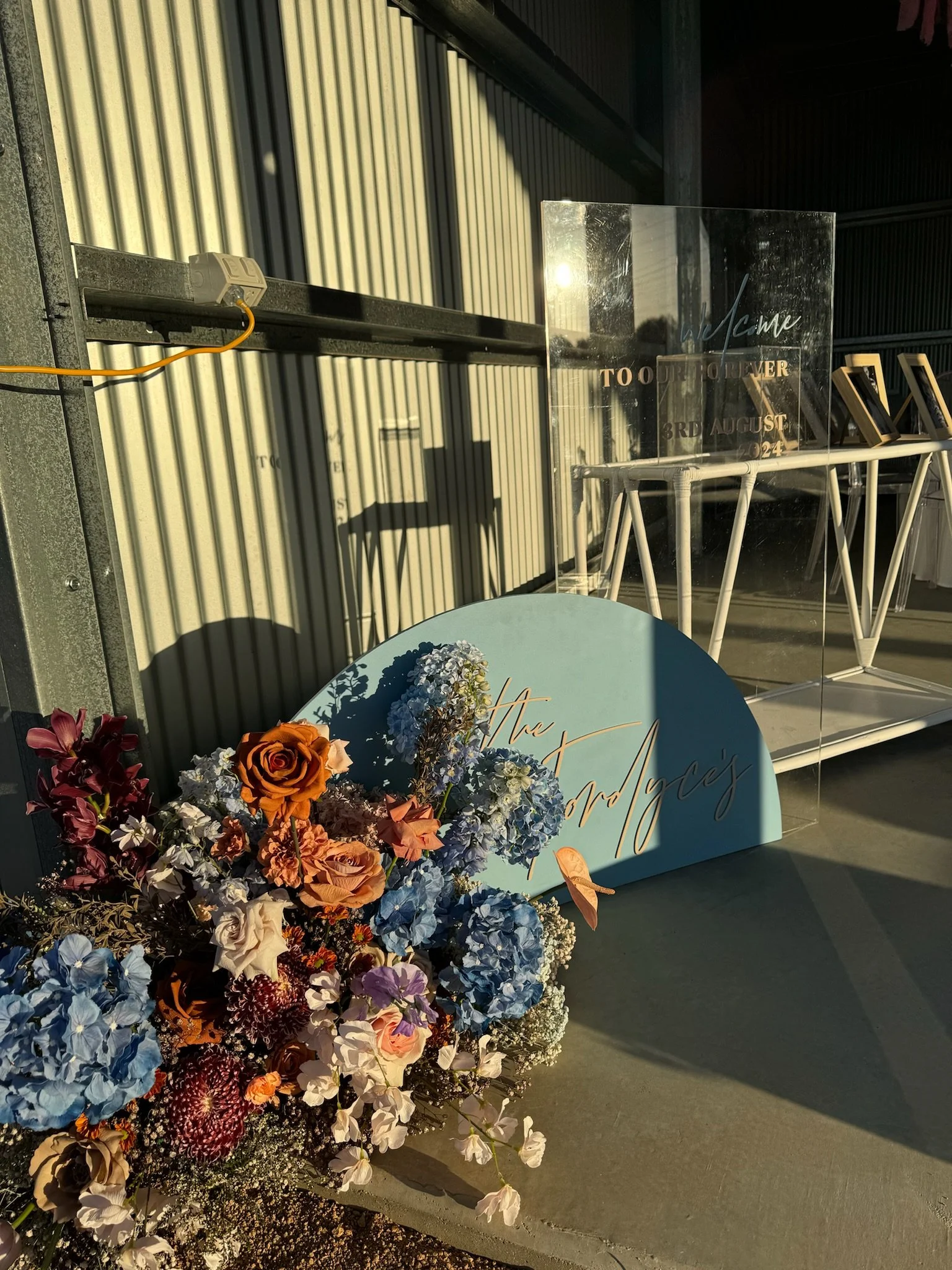 A floral arrangement with roses, hydrangeas, and other flowers in shades of blue, purple, white, and orange, placed next to a blue semi-circular sign with gold cursive writing that reads "The Adams". In the background, there's a transparent acrylic s