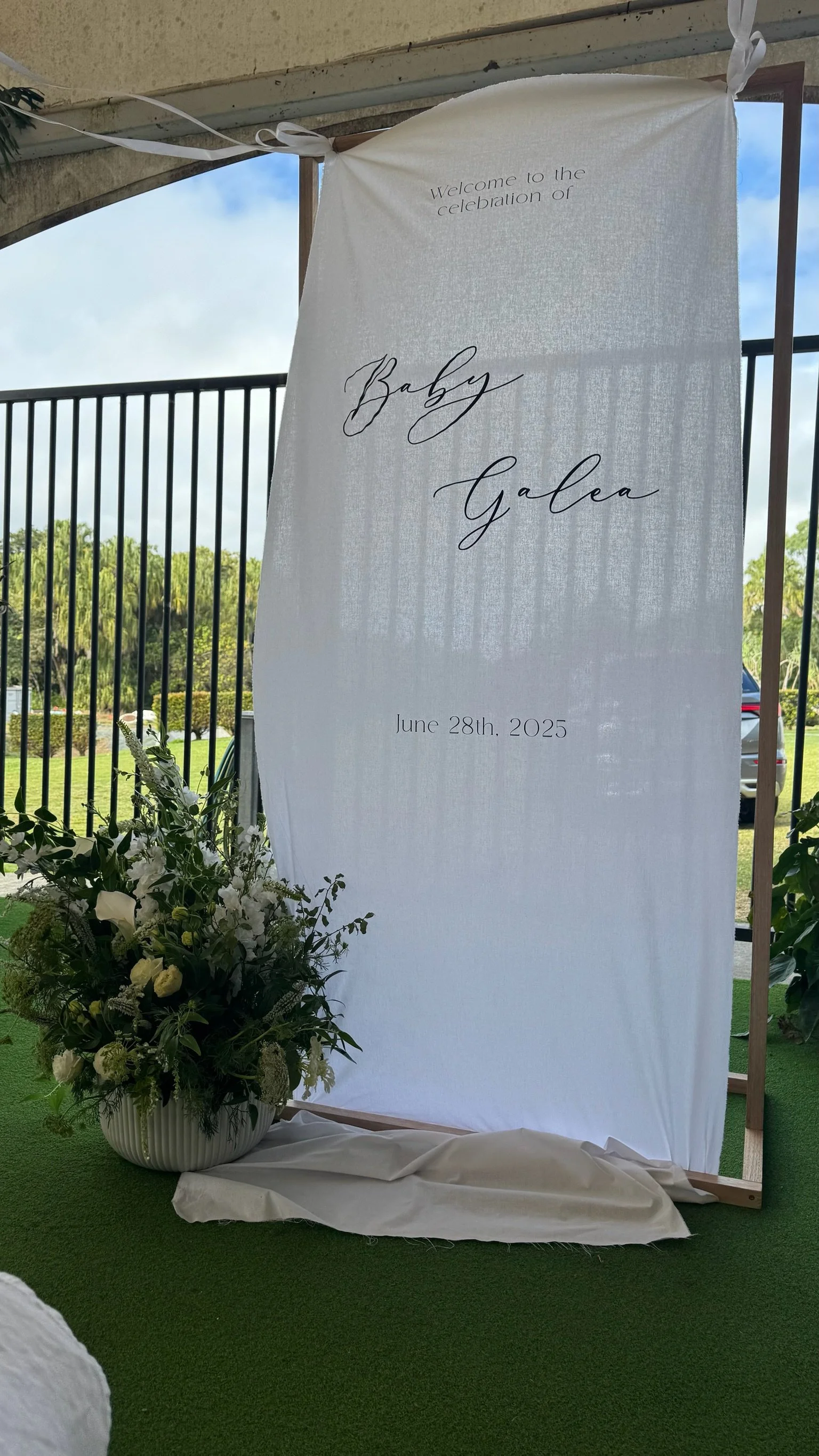 A white fabric banner with black cursive text that reads 'Welcome to the celebration of Baby Galea. June 28th, 2025'. It is attached to a wooden frame in an outdoor setting, with a floral arrangement of white flowers and greenery at the base. There i