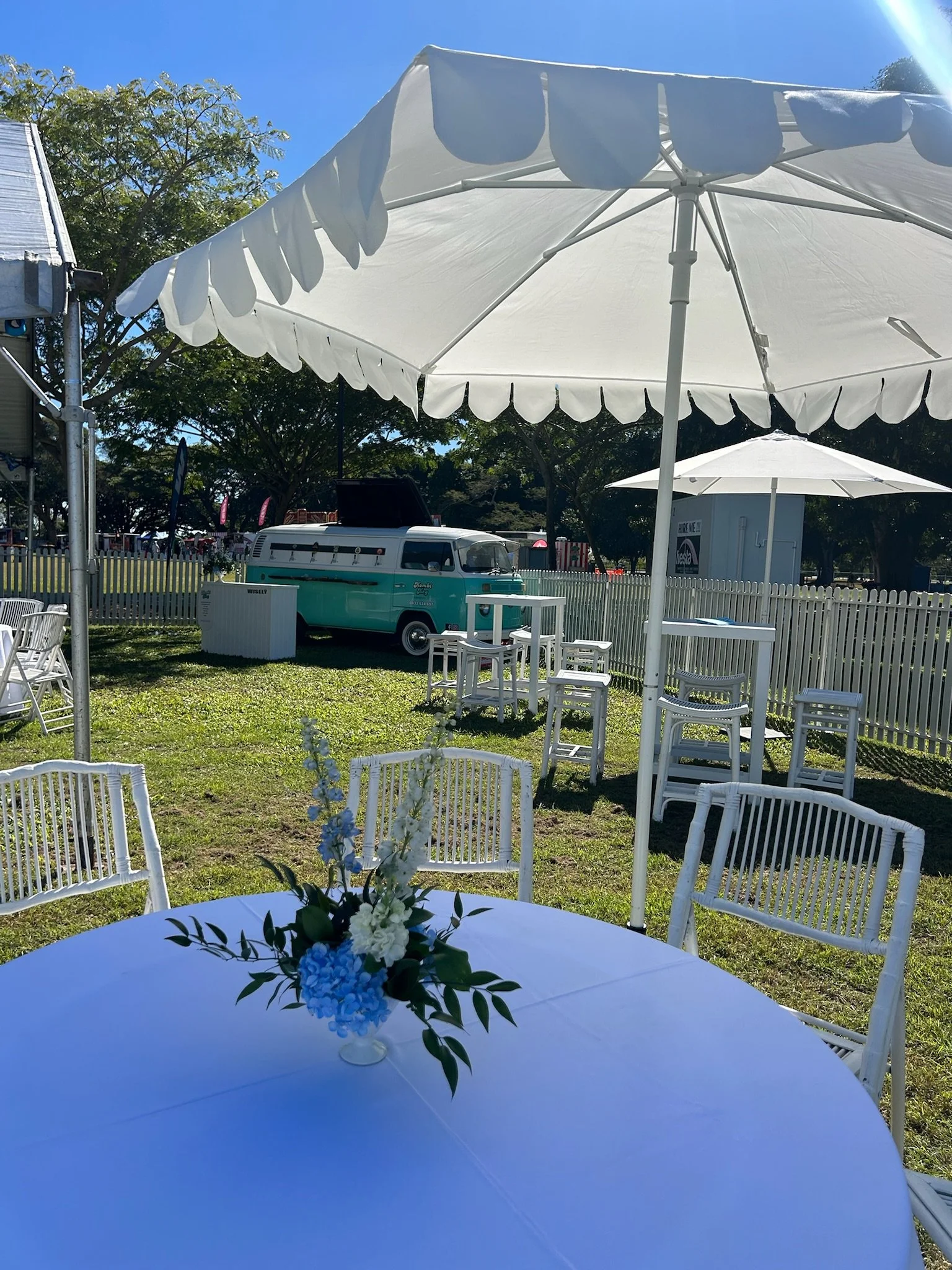 Outdoor event setup with white round table, white chairs, a centerpiece of blue and white flowers, large white umbrellas, a vintage teal and white van in the background, and a white picket fence, under a clear blue sky.