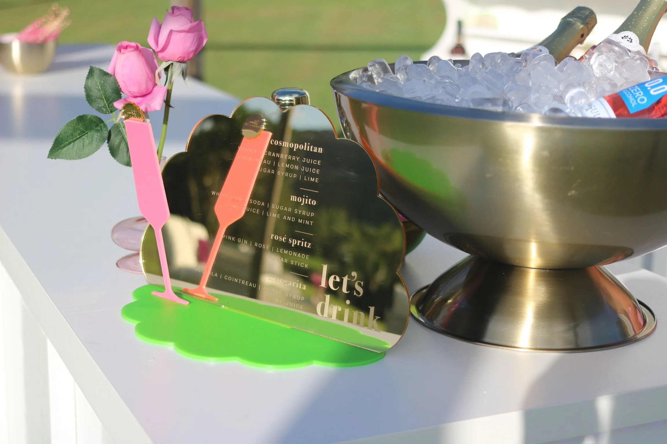 A table with a small pink rose bouquet, a reflective sign with cocktail names and ingredients, a green and pink drink stirrer, and a large metal bowl filled with ice and bottled drinks, outside on a sunny day.