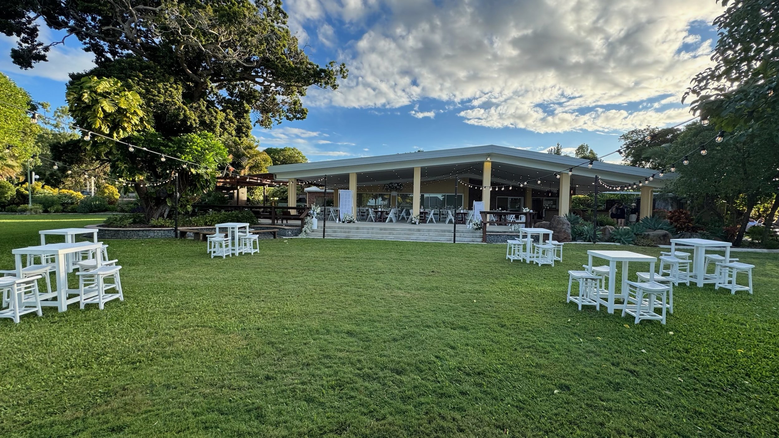 Outdoor event space with a large lush green lawn, several white tables with chairs, and a covered pavilion decorated with string lights under a partly cloudy sky.