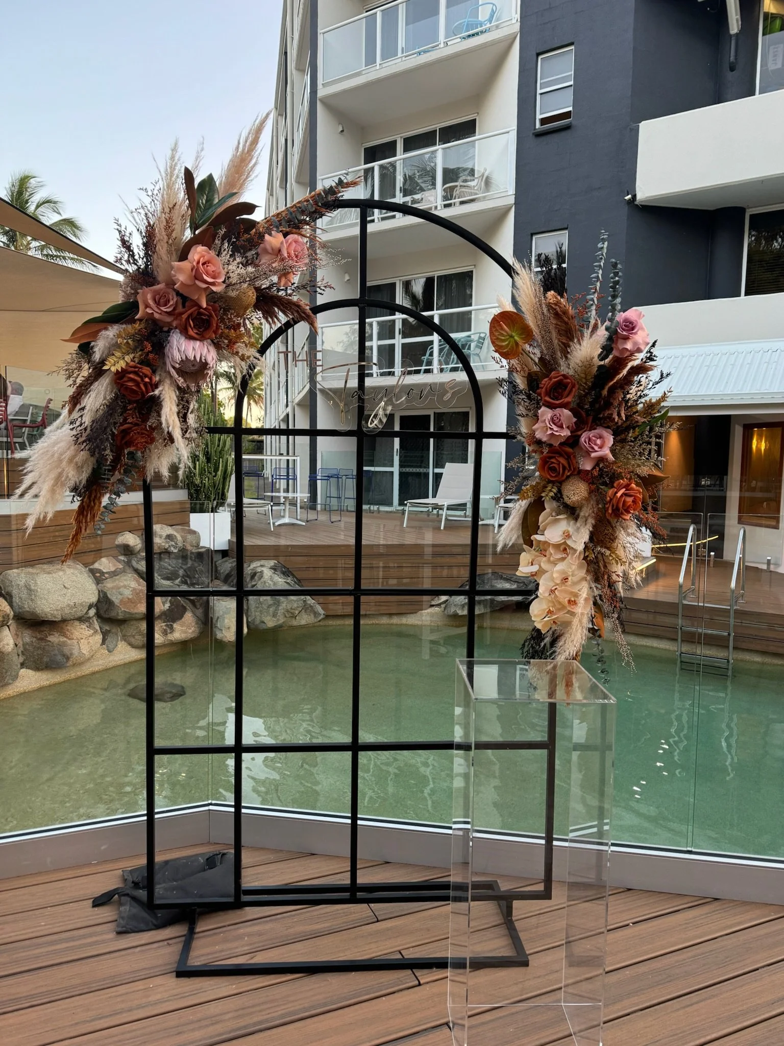 Decorative floral arrangement on a black metal frame, with pink and brown roses, pampas grass, and other dried flowers, on a wooden deck near a pool.