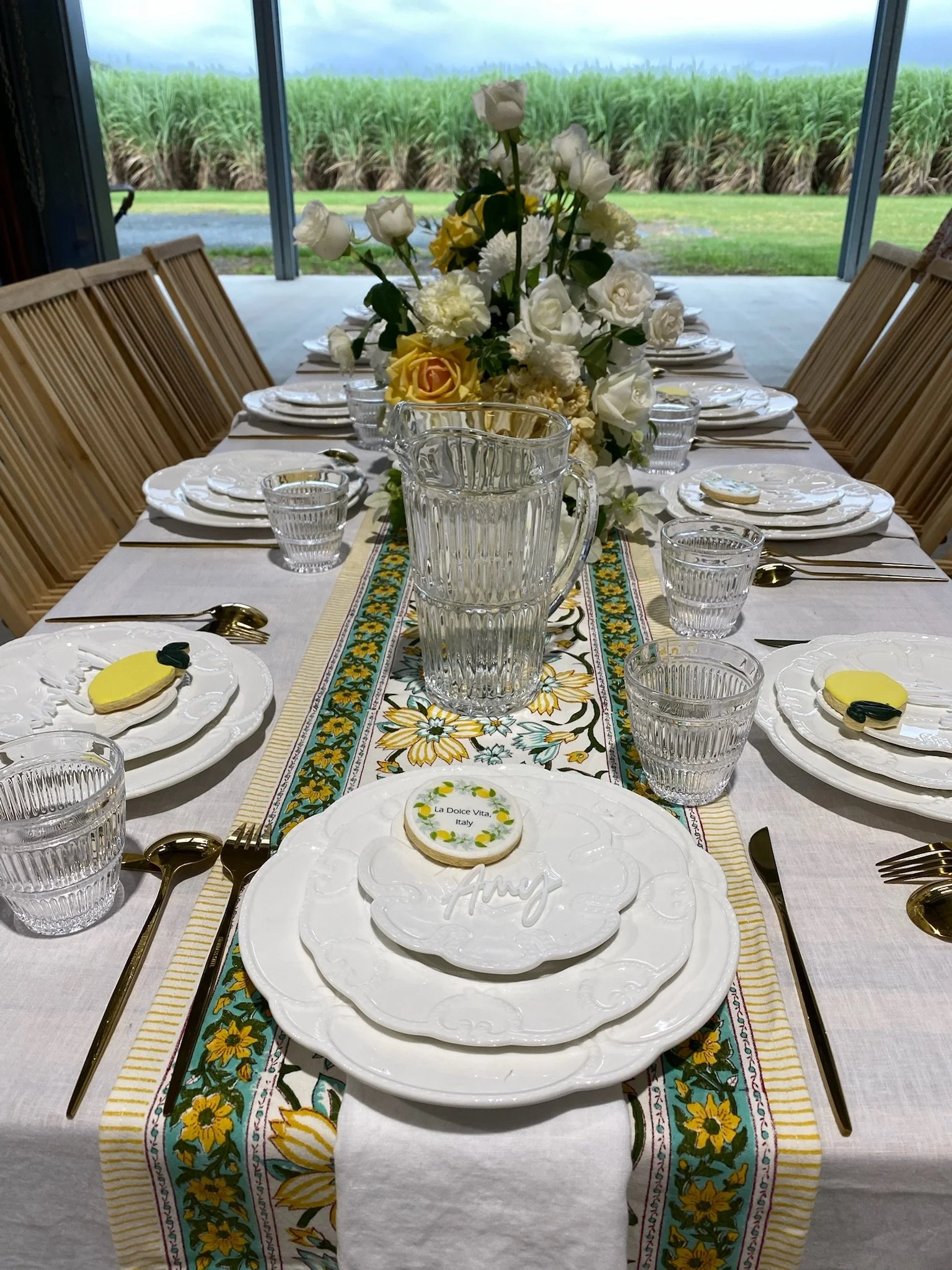 A dinner table set with white plates, gold utensils, and clear glassware, featuring a floral centerpiece with white roses and yellow flowers, with a scenic outdoor view of a green field and a barn in the background.