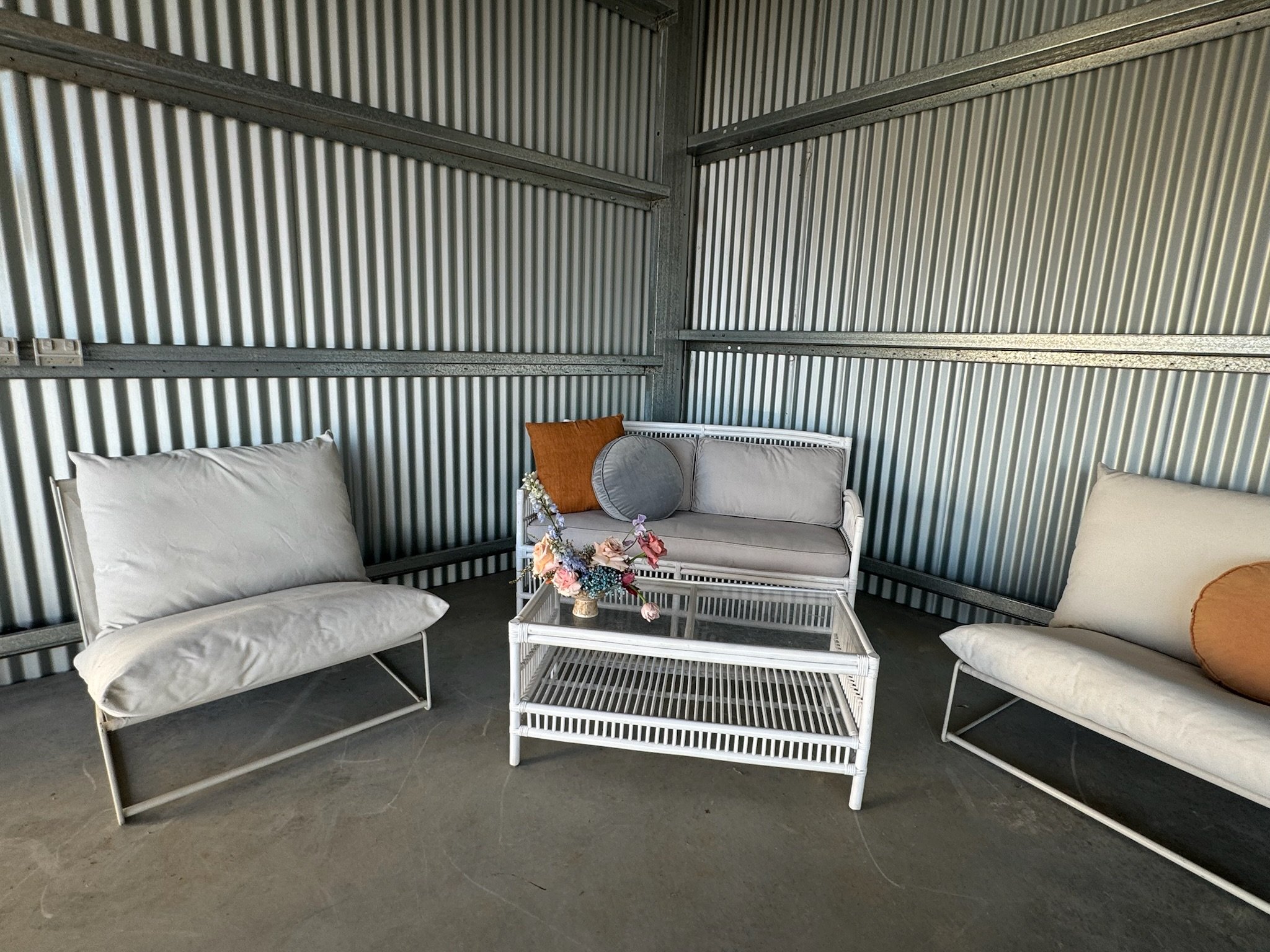 Empty indoor space with corrugated metal walls, white metal-framed furniture with light cushions, a glass-top coffee table with a flower arrangement, and decorative pillows.