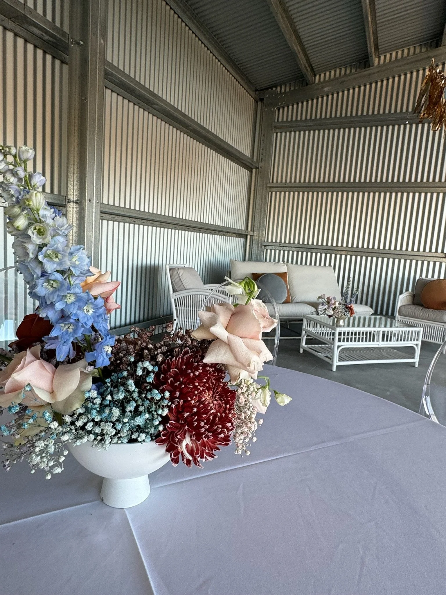 A table with a floral arrangement in a white vase, with white outdoor furniture including a sofa, chairs, and a coffee table in a covered outdoor area with corrugated metal walls.