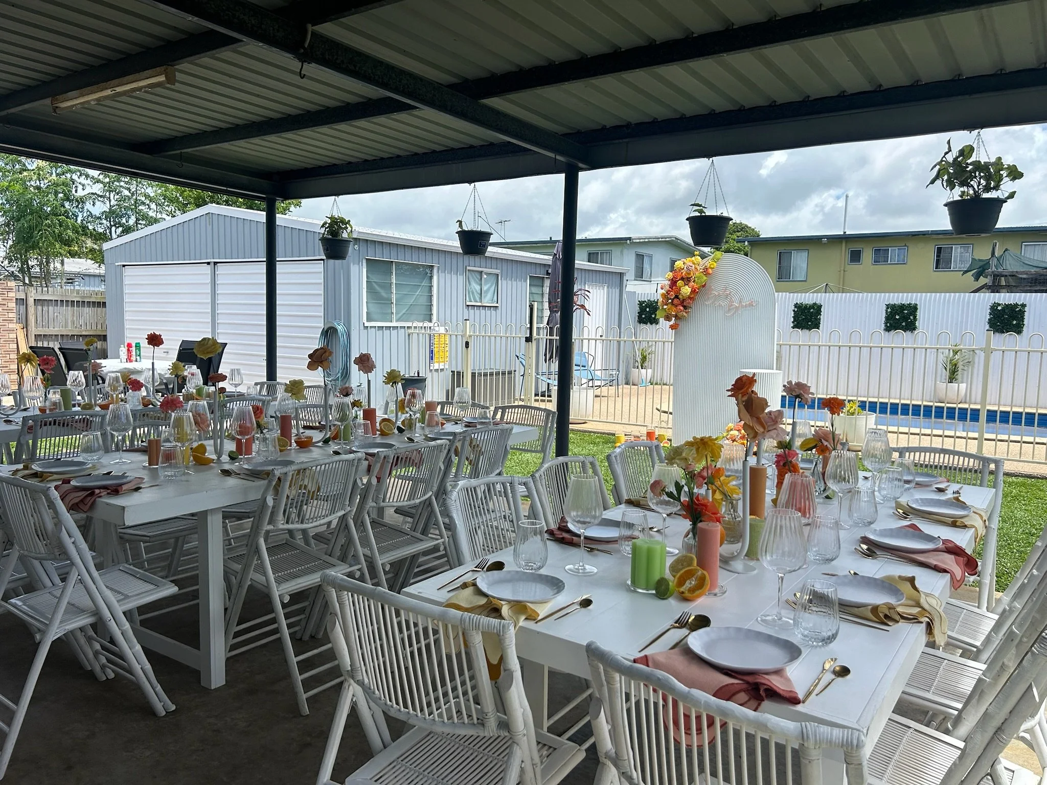 Outdoor party setup under a covered patio with white tables decorated with flowers, candles, and tableware, overlooking a backyard with a pool, white fence, and surrounding buildings.