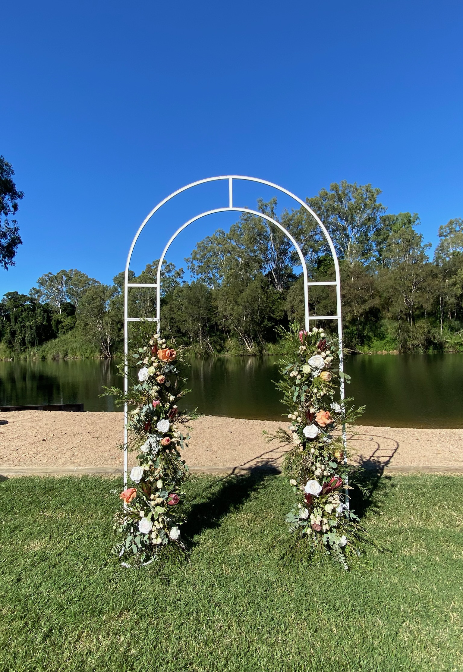 A white metal wedding arch decorated with floral arrangements on a grassy area overlooking a lake with trees in the background on a sunny day.