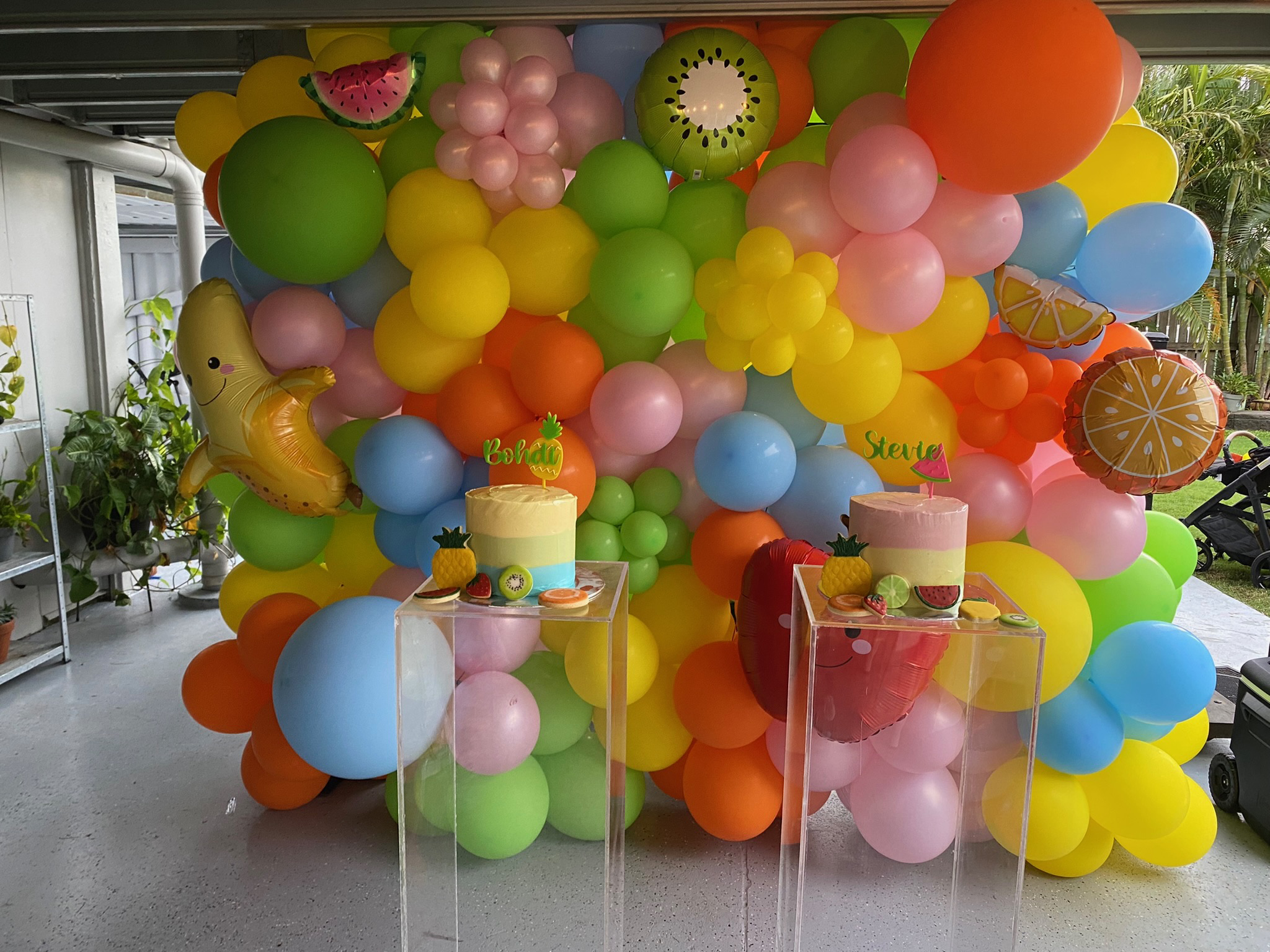 Colorful balloon backdrop with various fruit-themed balloons and two cakes on clear stands for a summer or tropical celebration.
