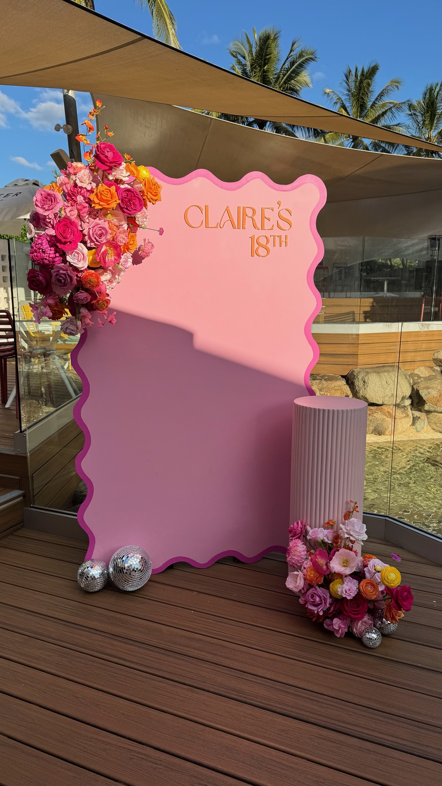 Pink birthday photo backdrop with floral arrangements on both sides, a white pleated lamp, and silver disco balls, celebrating Claire's 18th birthday on a wooden deck with clear glass sides and palm trees in the background.