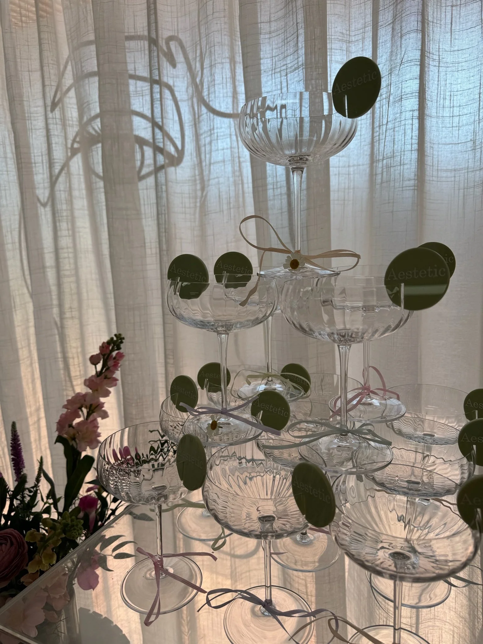 Glass champagne coupe glasses arranged in a pyramid on a transparent tiered stand, with tags that read "Aesthetic." The background features sheer curtains with light filtering through, and a bouquet of pink and purple flowers is visible on the left s