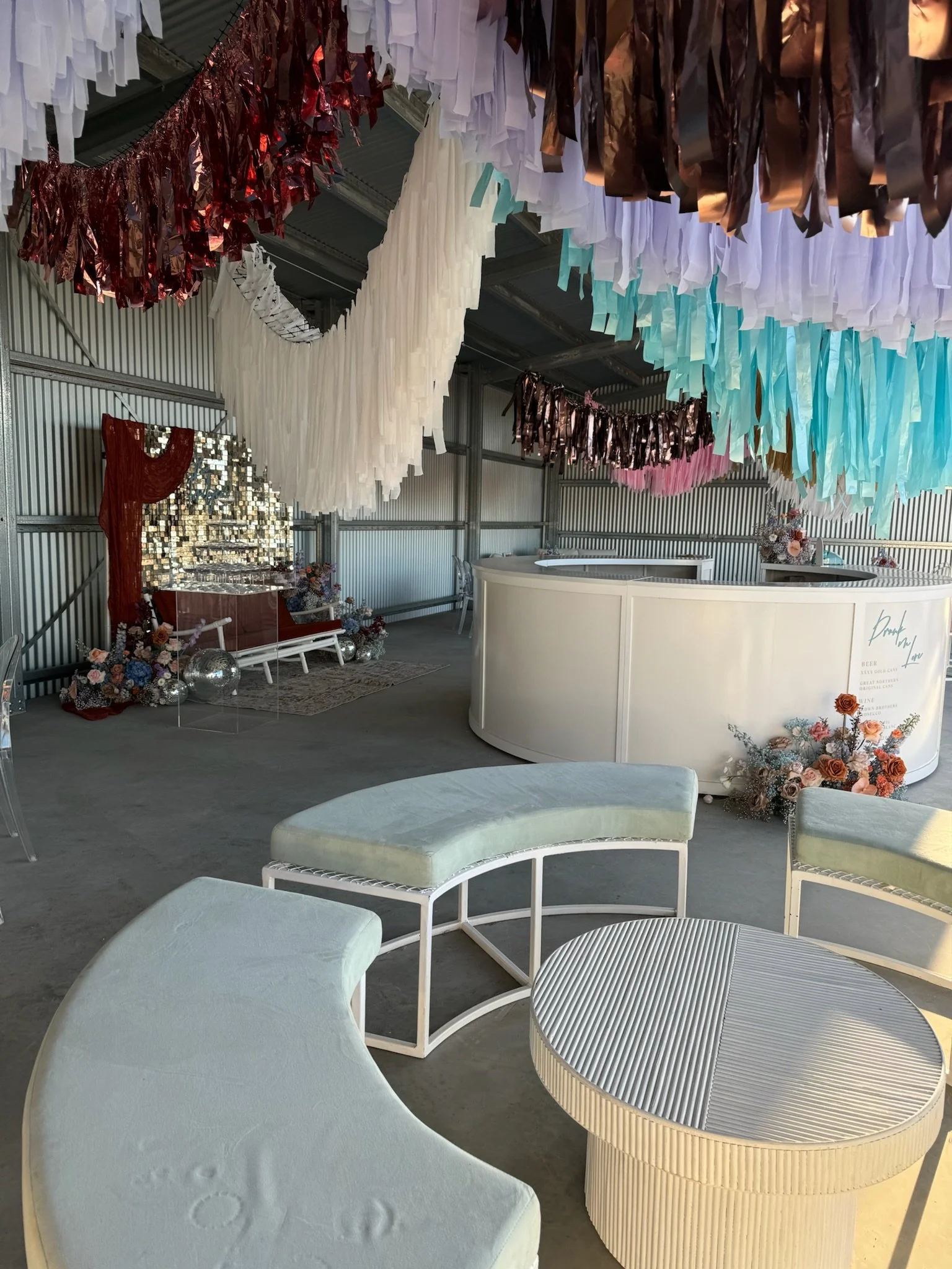 Colorful decorations hanging from the ceiling, including paper and metallic streamers, with a seating area featuring modern light-colored furniture and floral arrangements in the background.