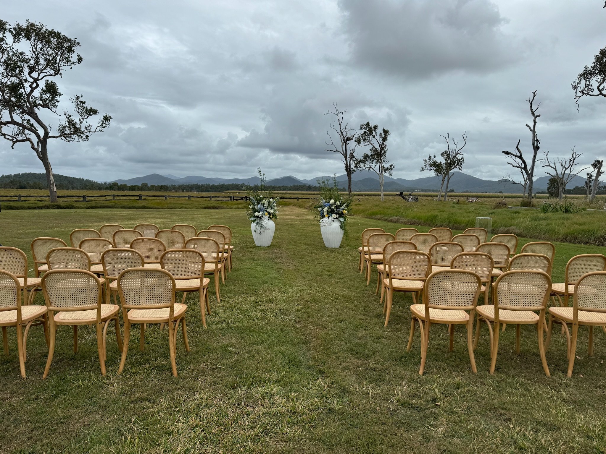 Outdoor wedding setup with chairs arranged on grass in a field with a cloudy sky, two large floral arrangements in white vases at the front, and barren trees in the distance, with mountains on the horizon.