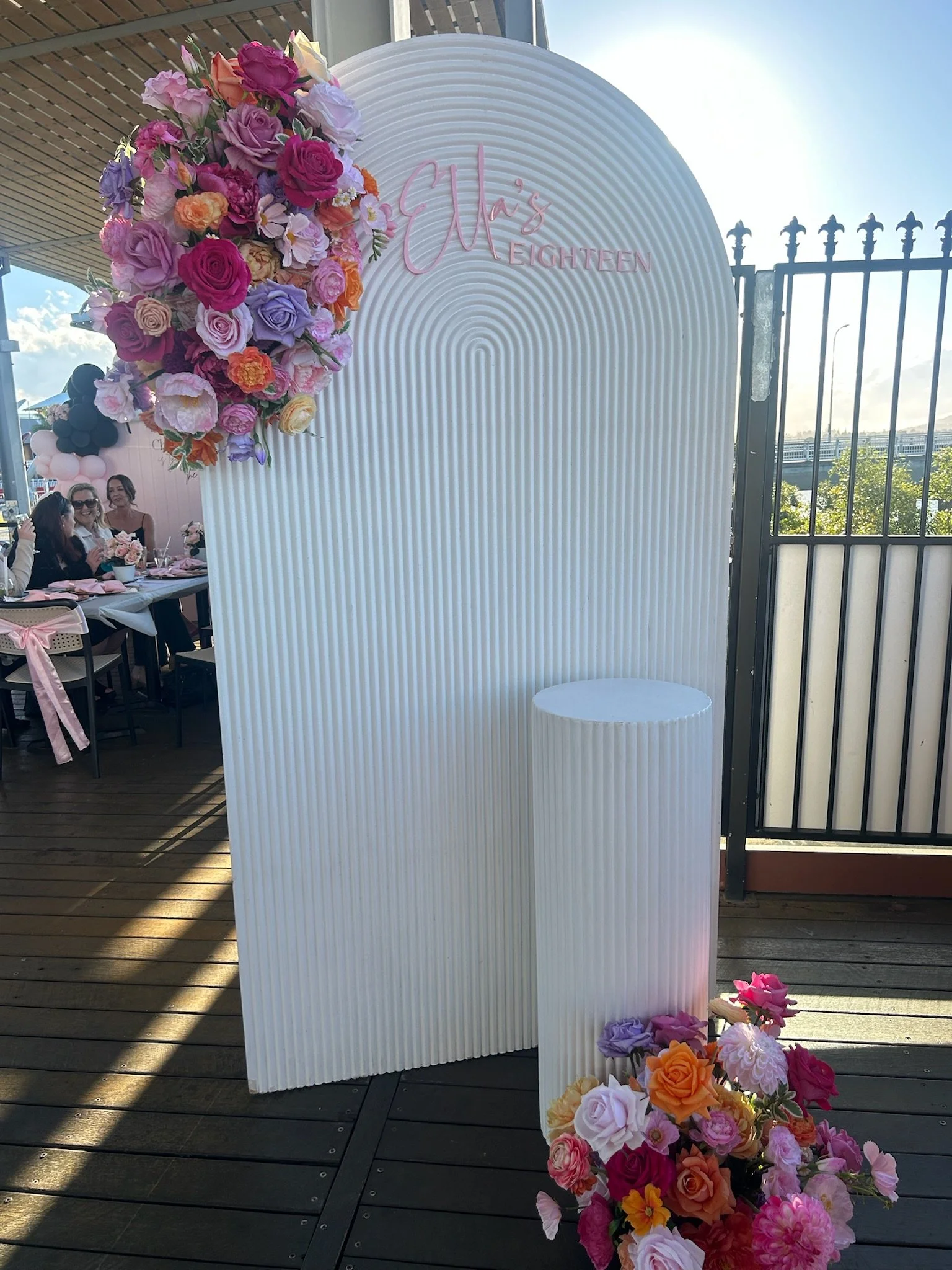 Decorative backdrop with floral arrangements and the message 'Elle's Eighteen' for a birthday celebration, with pink, purple, orange, and peach flowers, set on a wooden deck outdoors.
