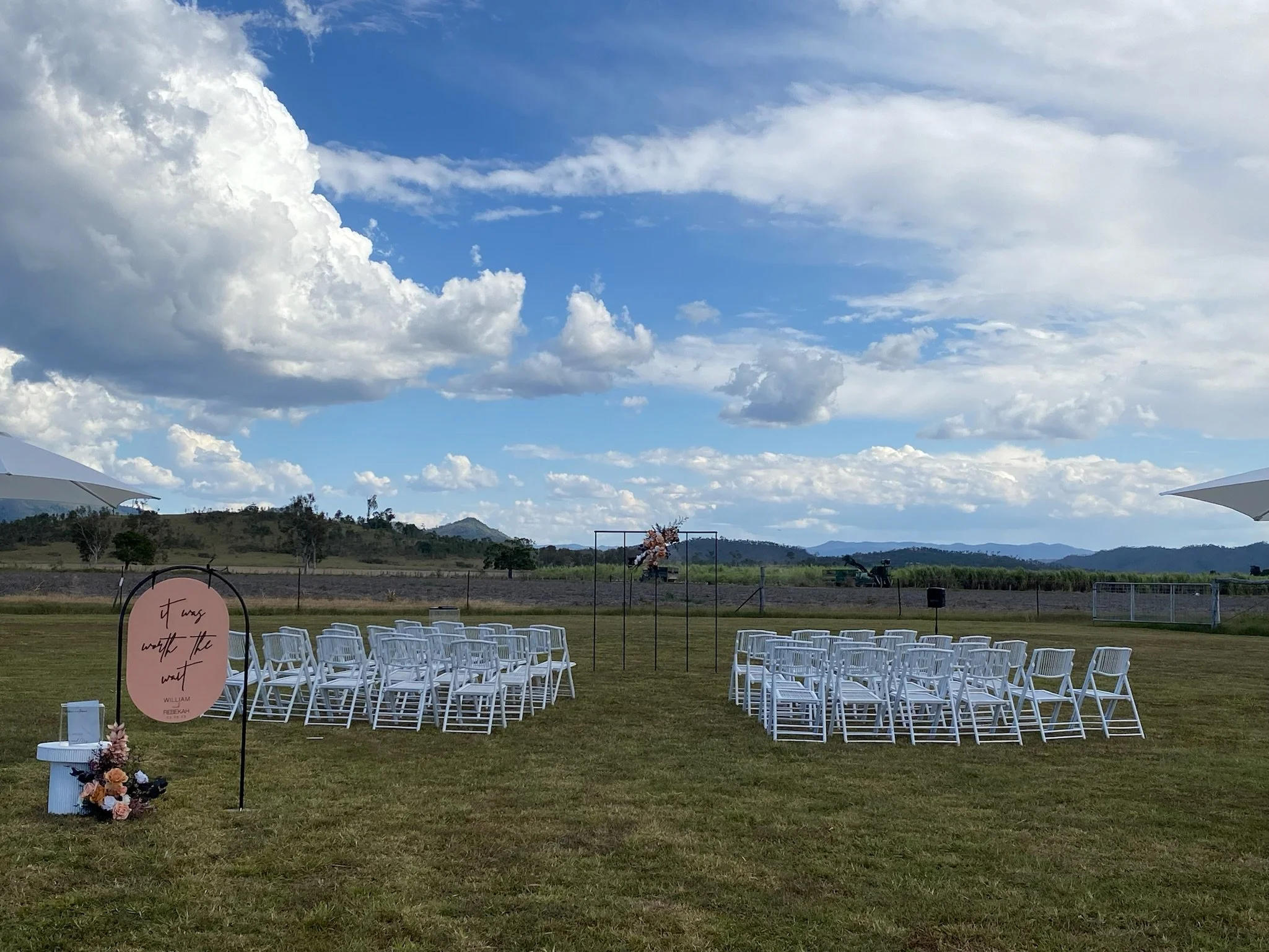 Outdoor wedding ceremony setup with white chairs arranged in rows on grass, a pink sign with text, floral decorations, and umbrellas on a sunny day with clouds and mountains in the background.