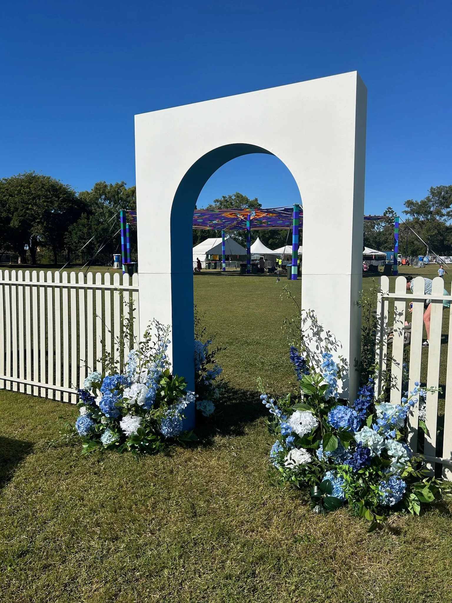 White decorative archway with floral arrangements of white and blue flowers at its base, set on a lawn during a sunny day.