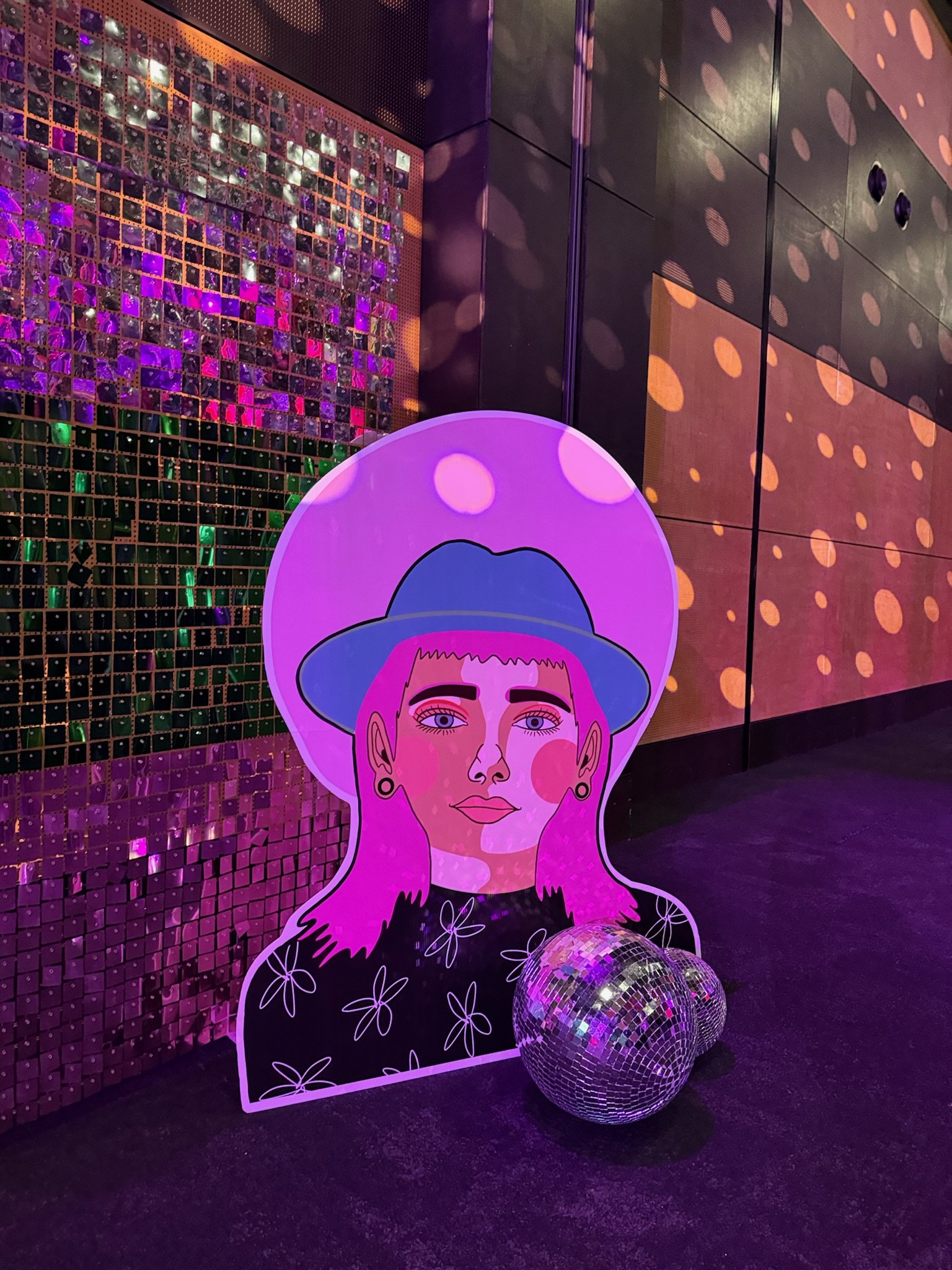 A colorful, illuminated artwork of a woman with pink hair, wearing a blue hat, with a disco ball at her feet, set against a backdrop of shiny, reflective tiles and dotted lights.