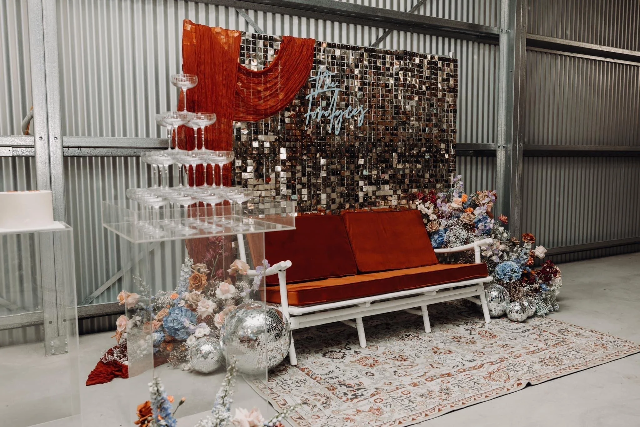 A decorative setup featuring a white bench with orange cushions, surrounded by flowers, disco balls, and a backdrop with a mosaic pattern and a sign that reads 'The Fortneys.' There is a clear acrylic tiered stand with empty wine glasses on the left 