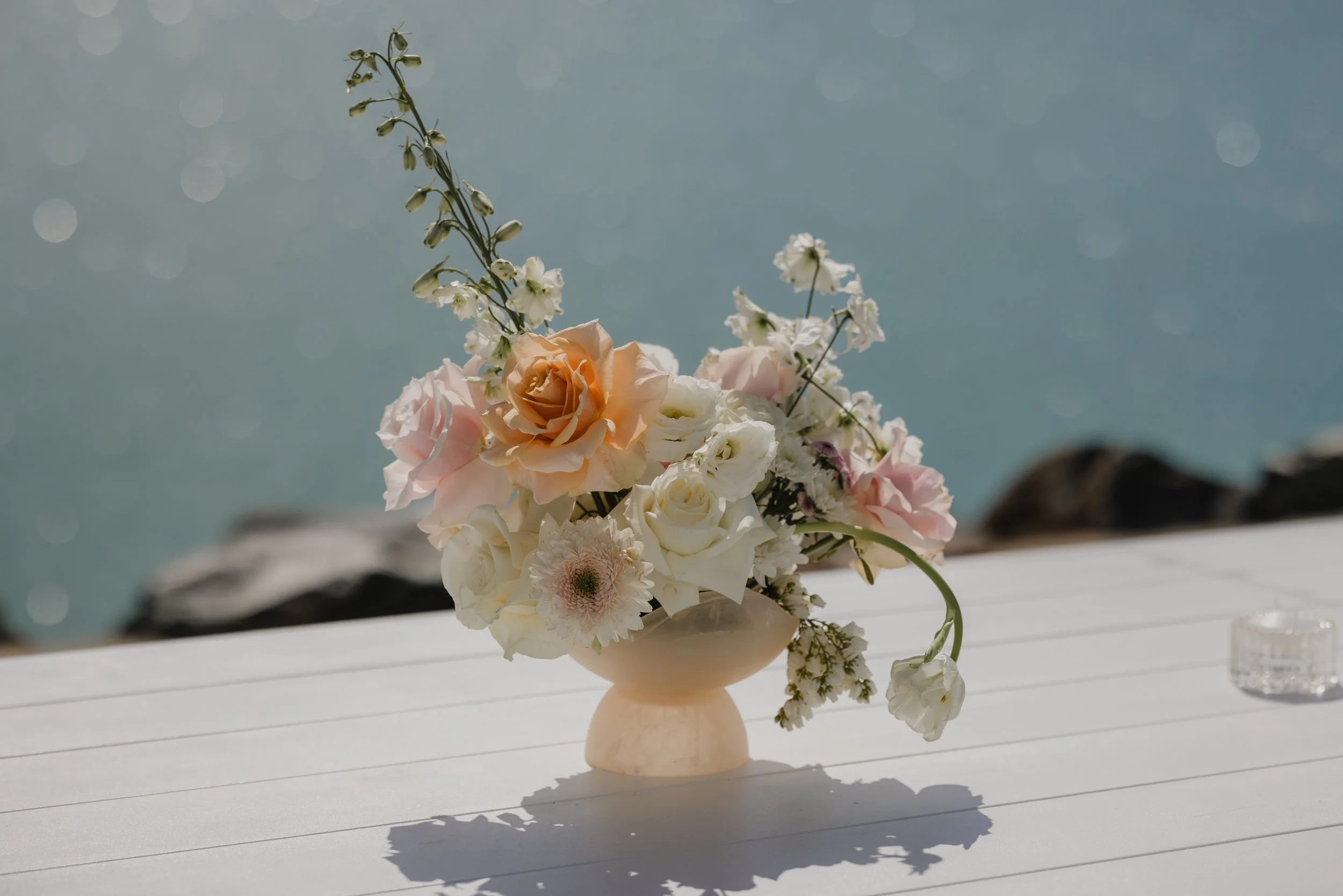 A bouquet of various pastel-colored flowers in a cream-colored vase on a white table, with a body of water in the background.