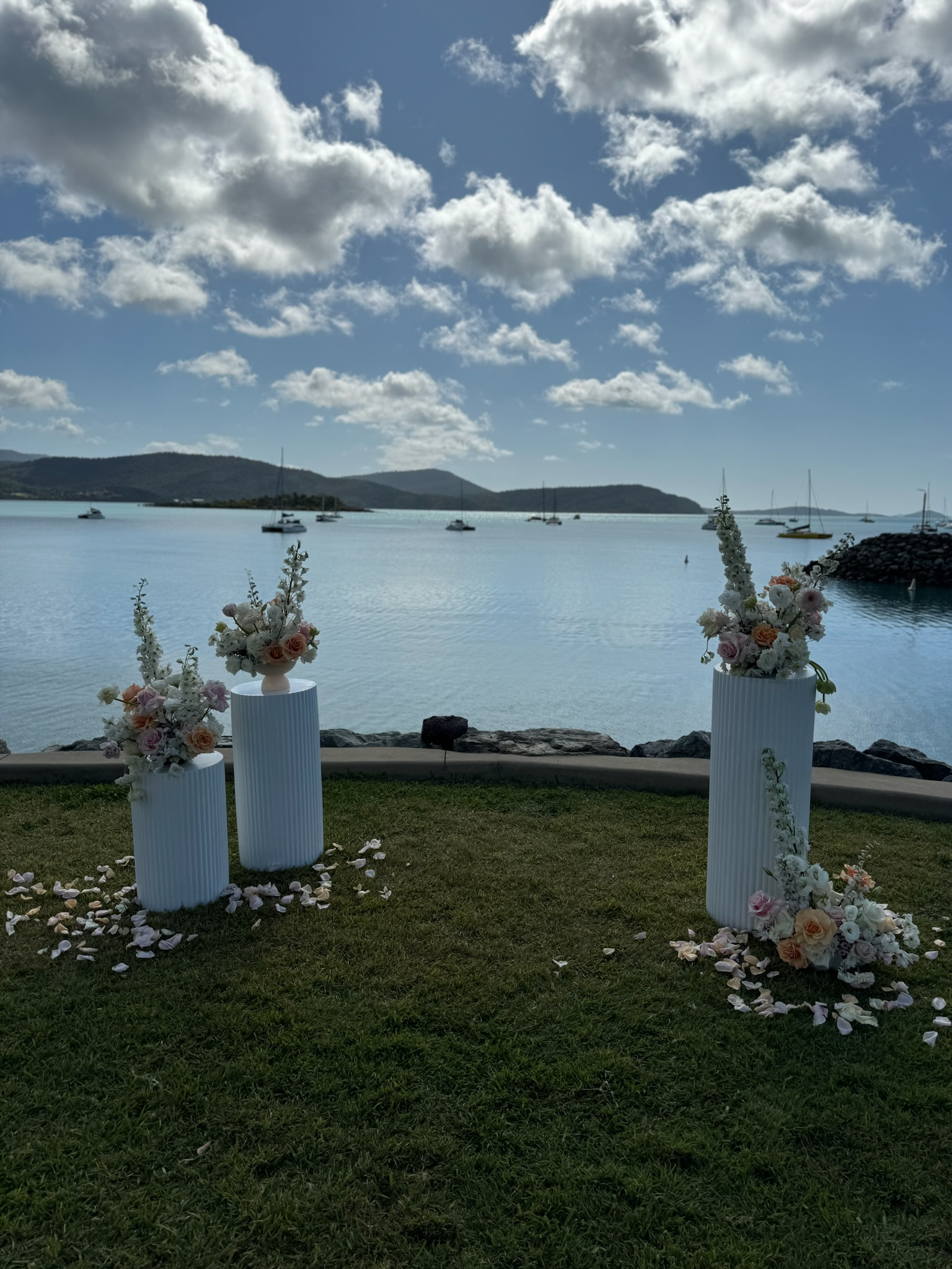 Decorative floral arrangements on white pedestal vases set outdoors near a body of water with sailboats, on a grassy area with scattered flower petals, under a partly cloudy sky.