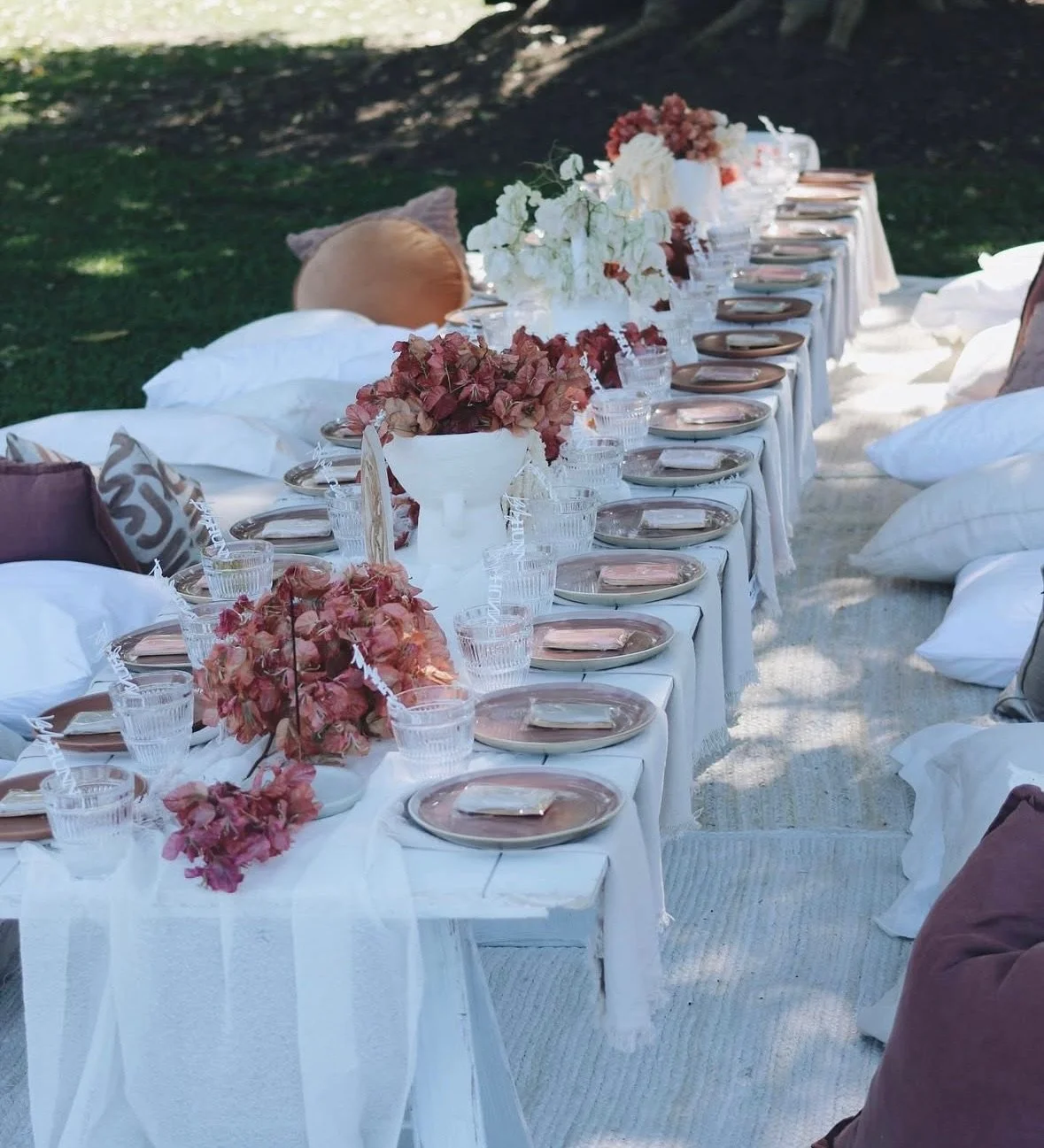 Outdoor dining table set for an event with pink and white floral centerpieces, pink plates, clear glasses, and pillows placed around it.