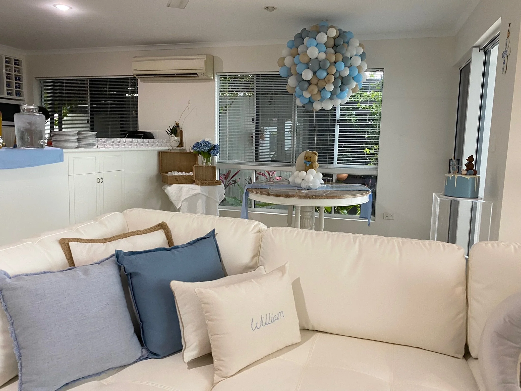 Living room decorated for a celebration with a white sofa, blue and white pillows, a round table with a blue and white balloon arrangement, and a cake with teddy bear toppers.