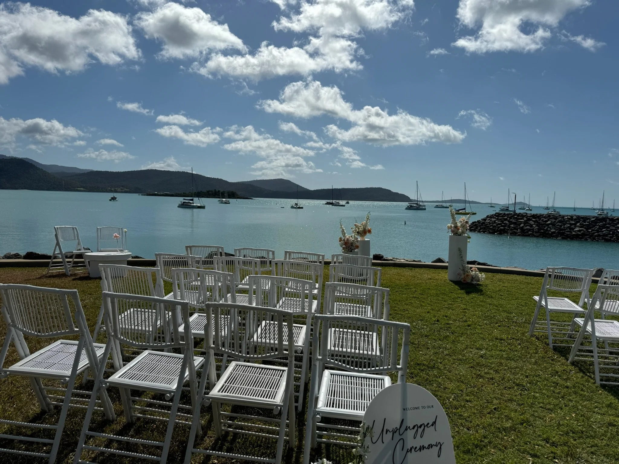 Outdoor wedding ceremony setup by the water with white chairs, floral arrangements, and a scenic view of sailing boats on the lake under a partly cloudy sky.