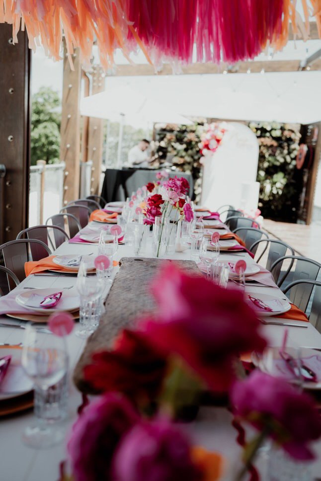 Long table set for a celebration with pink and orange floral arrangements, plates, glasses, and pink accents under colorful hanging decorations.