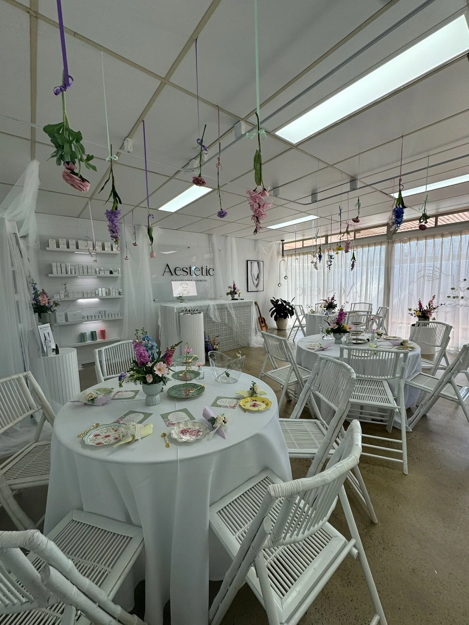 A beautifully decorated indoor event space with white tables and chairs, floral centerpieces, hanging pink and purple flowers, and a sign that reads 'Aesthetic' on the back wall.