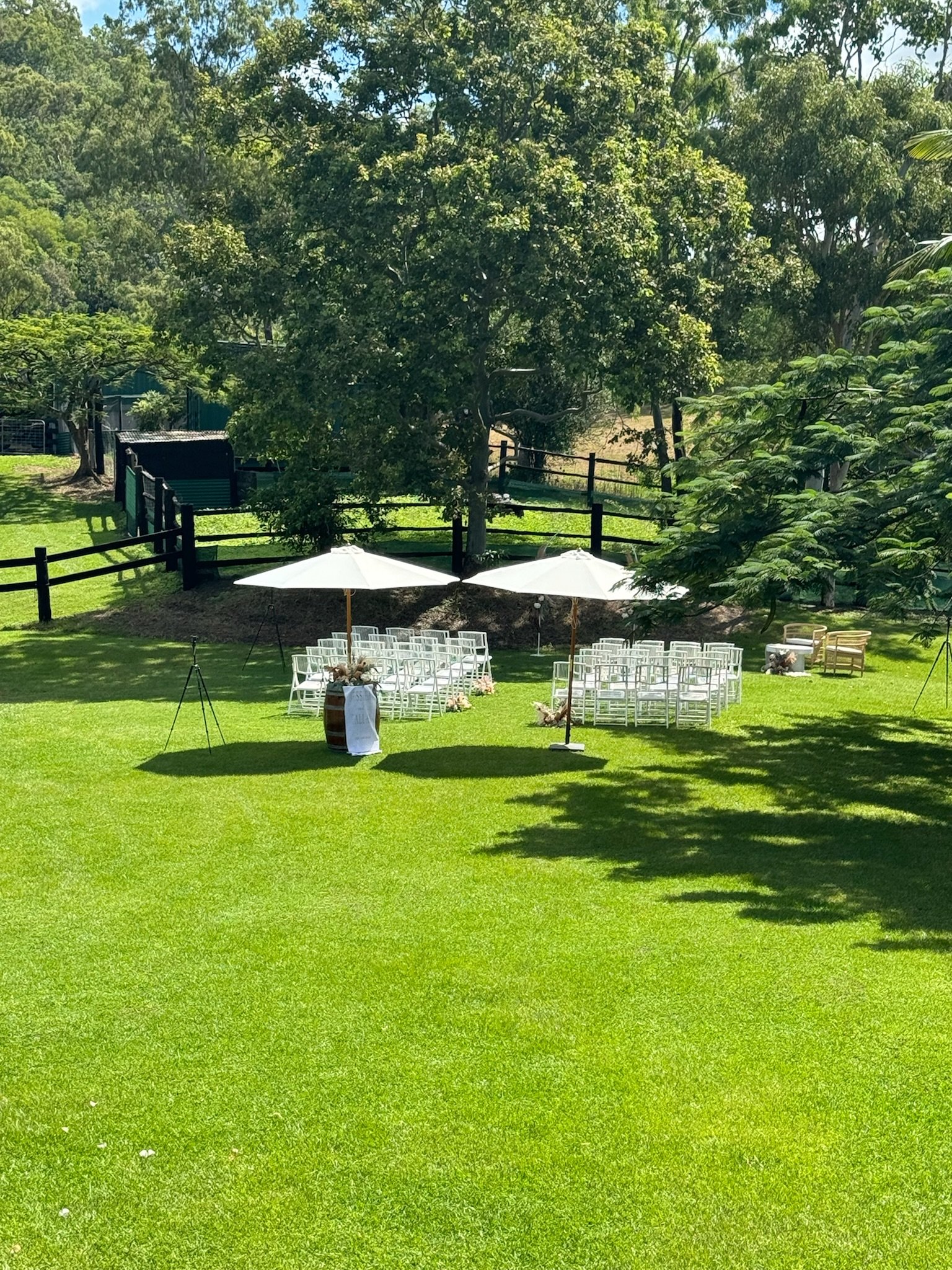 Outdoor wedding ceremony setup with white chairs arranged in rows, white umbrellas, decorative floral arrangements, and trees in a lush green yard.