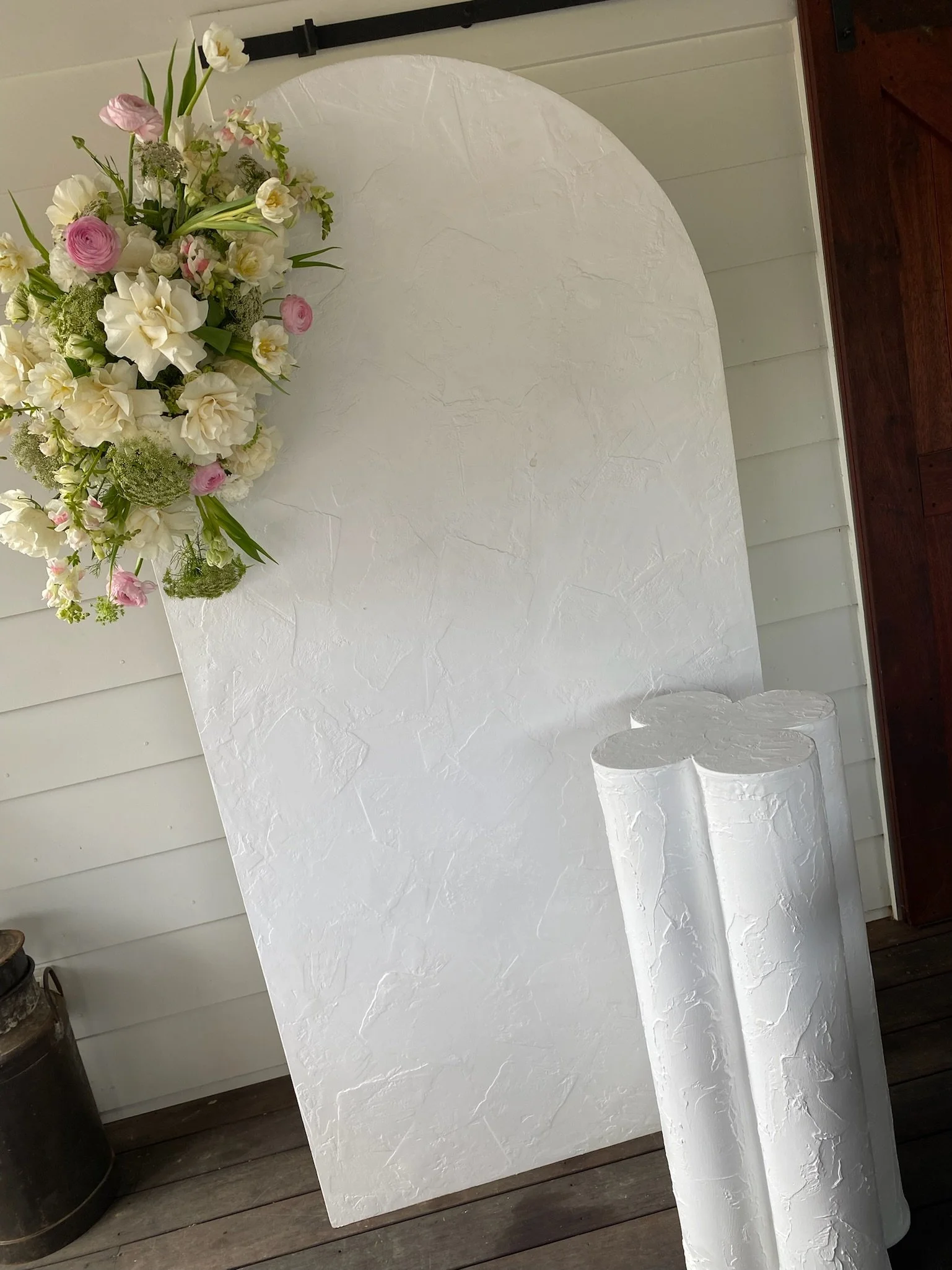 A decorative backdrop with a large white textured board, decorated with a floral arrangement of white and pink flowers, and three white textured cylindrical pillars in front.