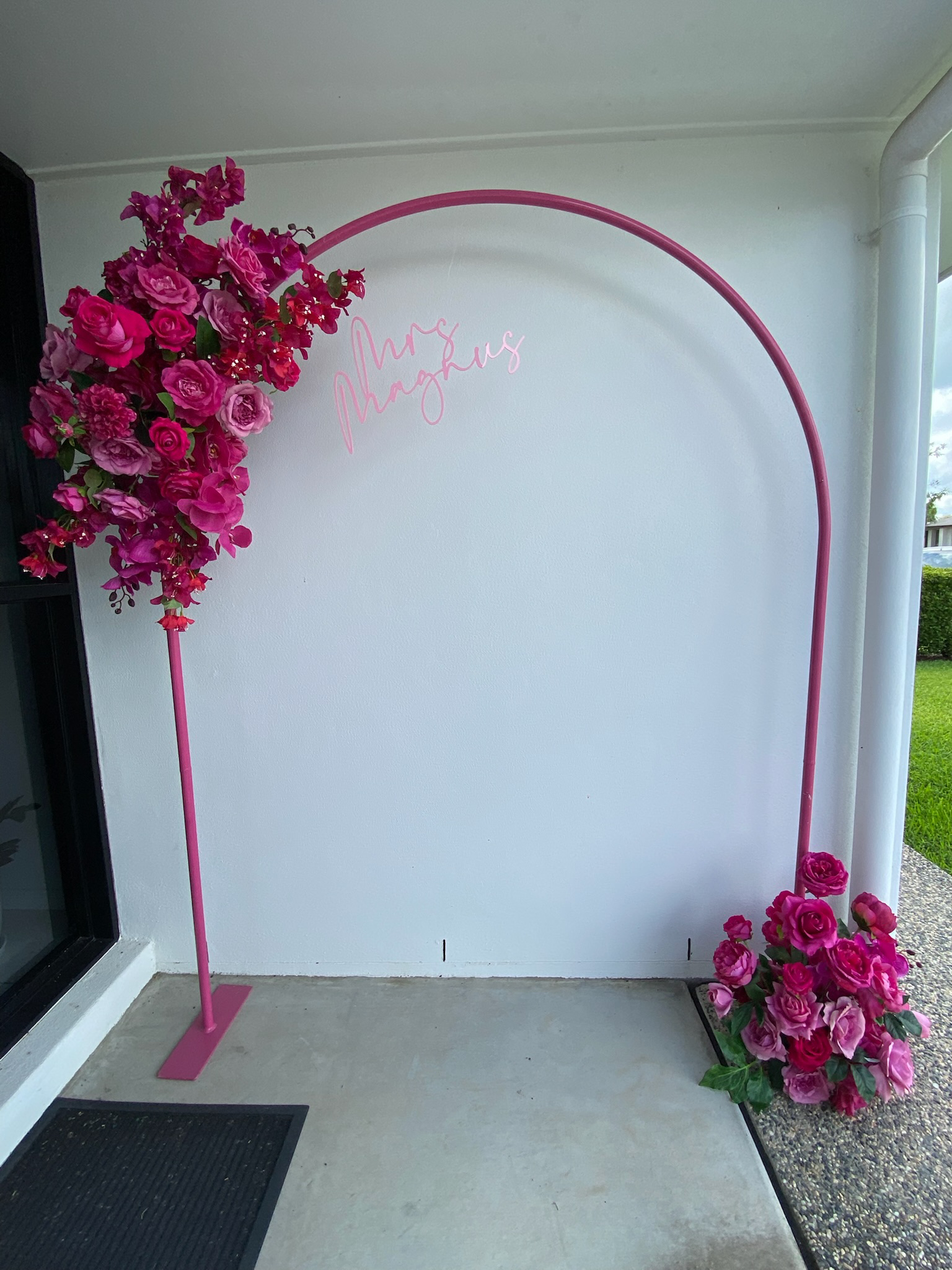 Pink floral arch with a neon sign that says 'Mrs. Manguis' on a white wall outside.
