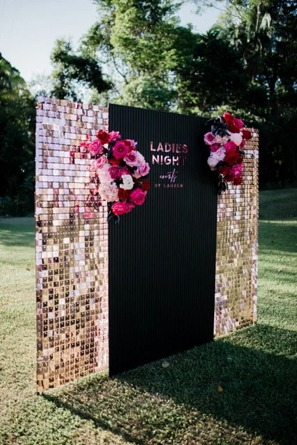 A decorative outdoor photo backdrop with a black center panel reading "Ladies Night" and floral arrangements on each side, surrounded by a mirrored mosaic frame on a grassy area with trees in the background.