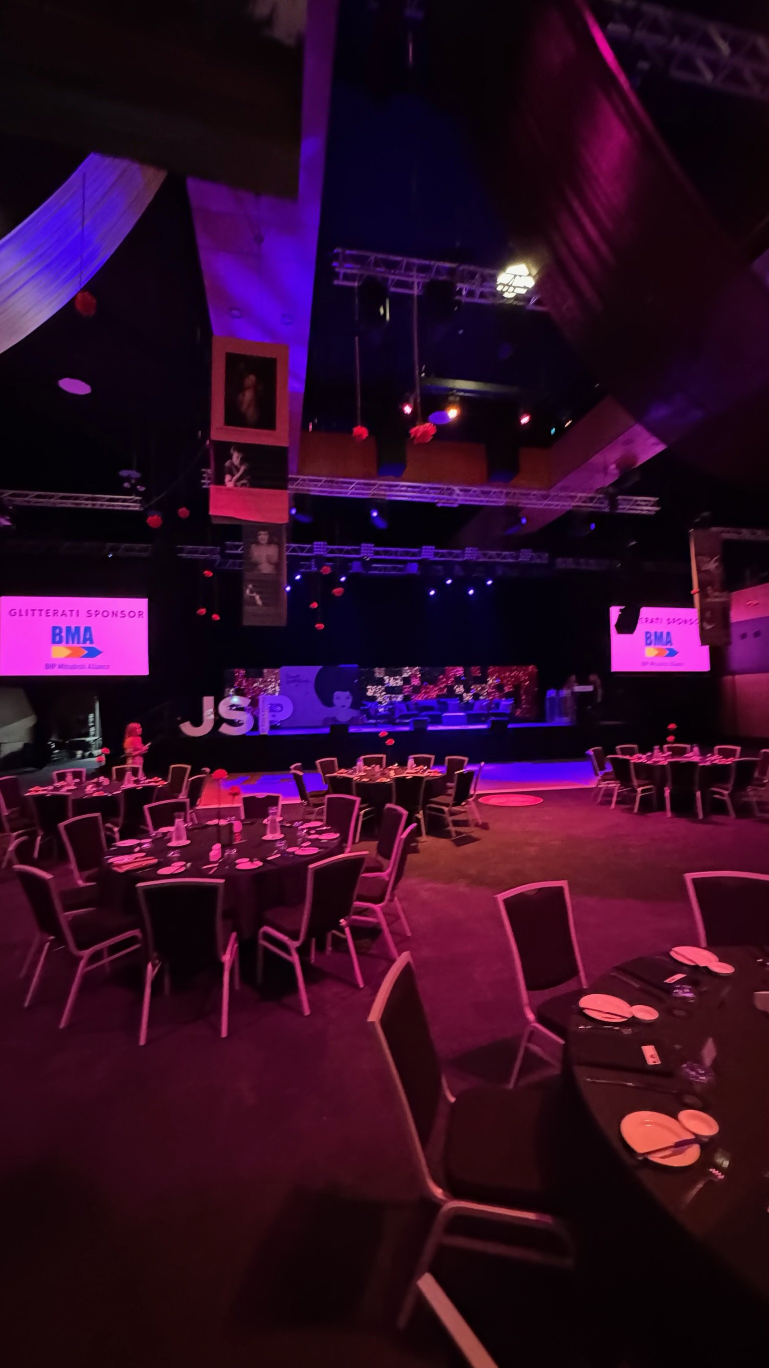 Event venue set with round tables and chairs, stage with screens and decorations, dim lighting with pink and purple hues.
