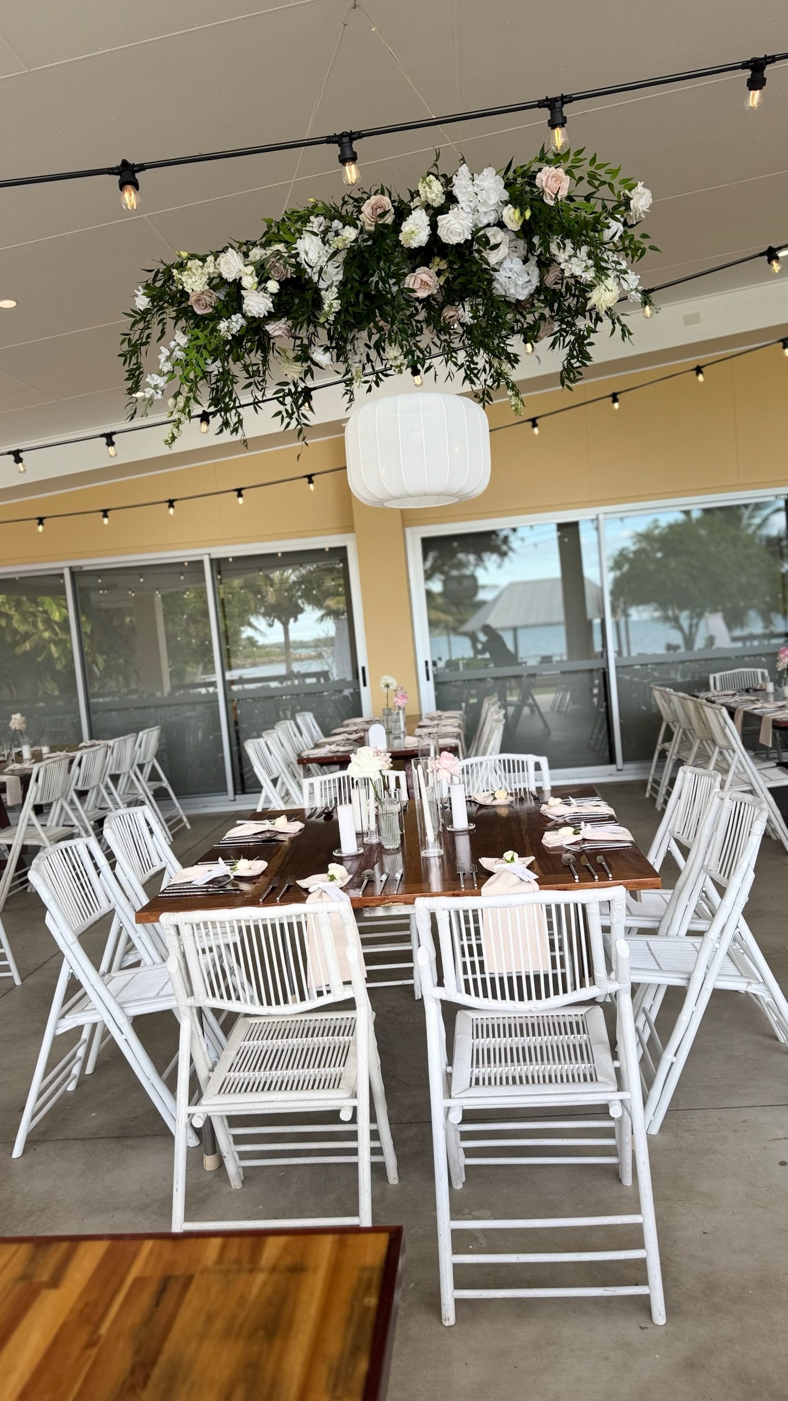 A decorated outdoor dining area with a hanging floral arrangement, string lights, and a table set with white napkins, candles, and tableware, overlooking a waterfront view through large glass sliding doors.