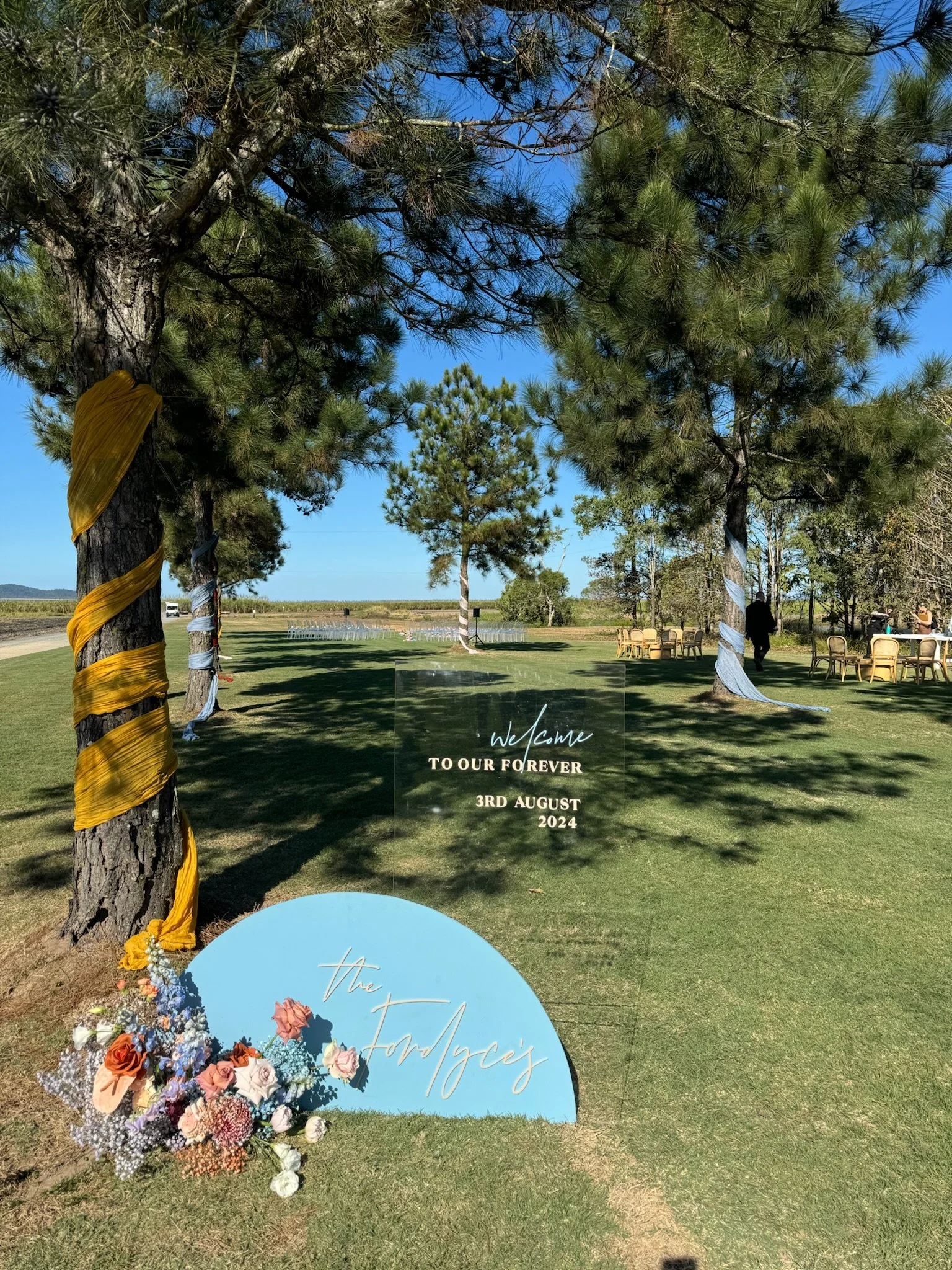 Outdoor wedding decoration with trees wrapped in fabric, a sign with wedding details, and floral arrangements on the grass.