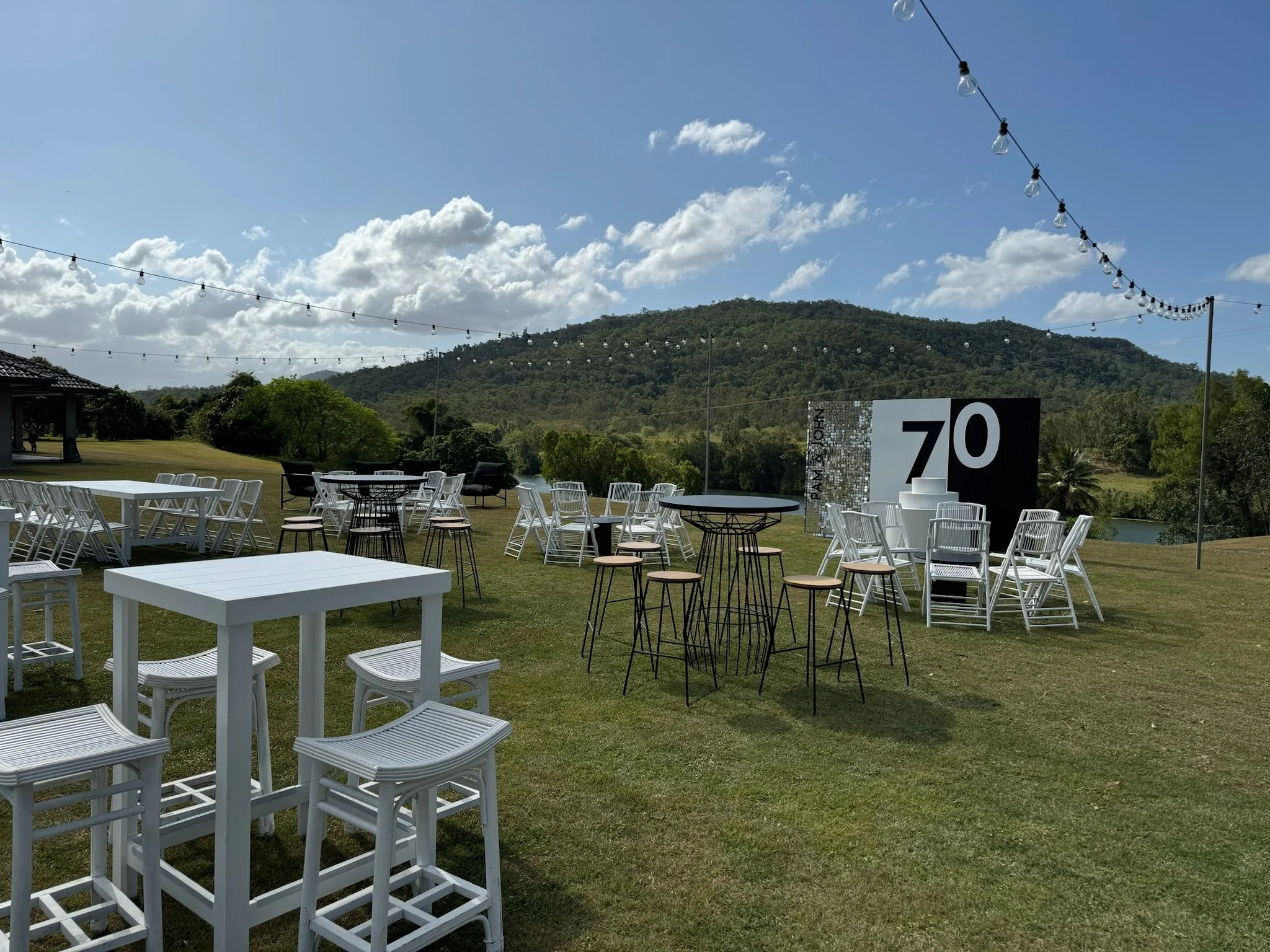 Outdoor event setup with white tables and chairs, high tables with stools, string lights overhead, a large painting with '70' in black and white, grassy area, hills and trees in the background, under a partly cloudy sky.