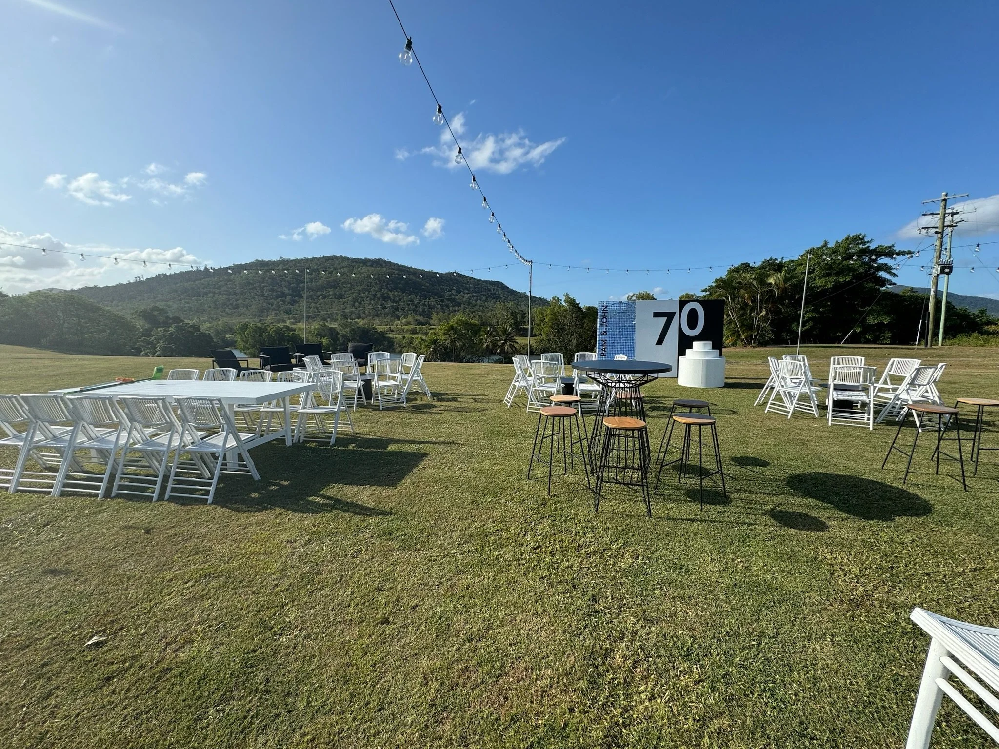 Outdoor event setup with white chairs and tables on a grassy field, with hills and a blue sky in the background. String lights are hanging overhead. There is a black round table with stools, and a large sign with the number 70.