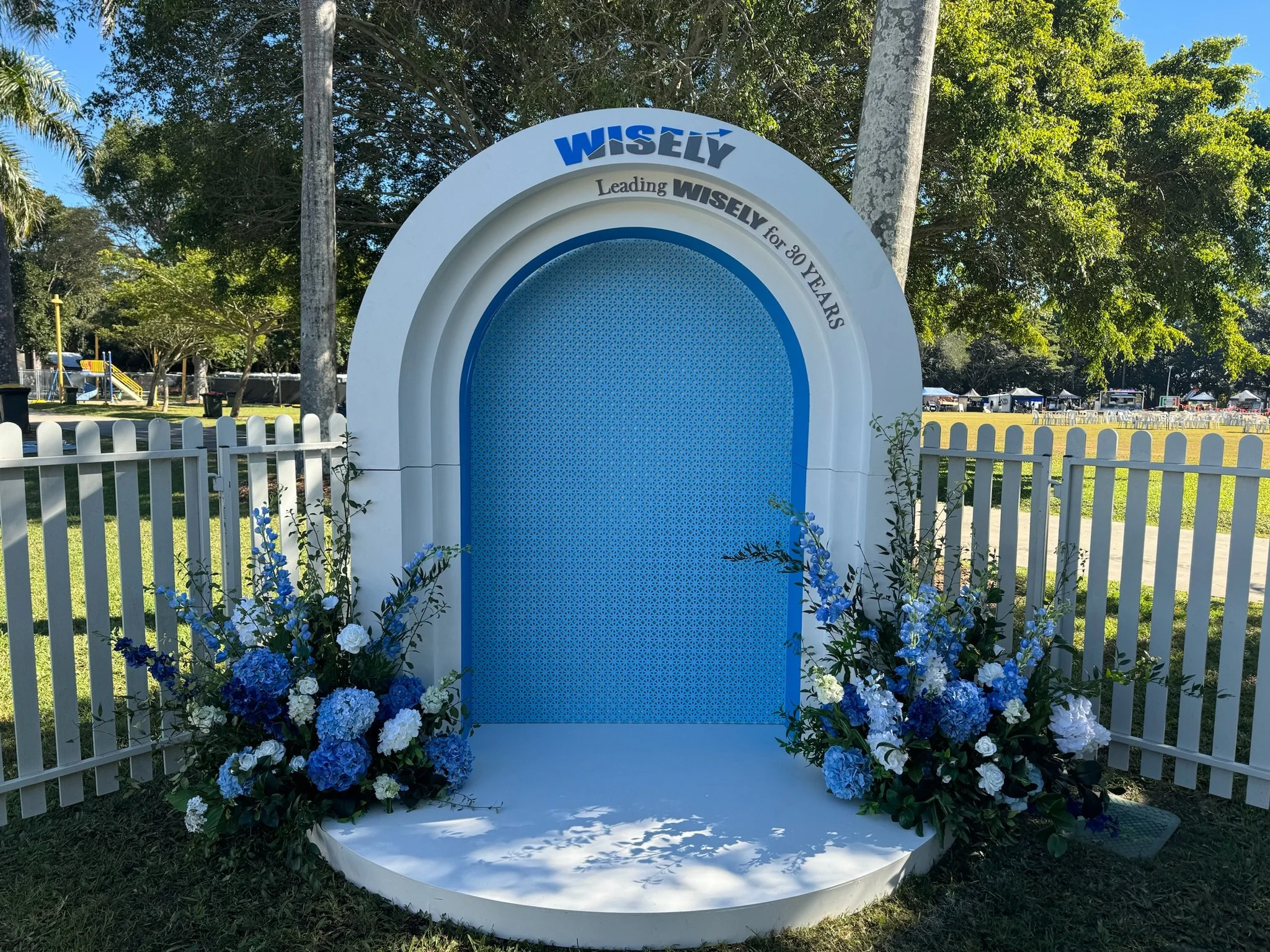 A decorative backdrop with white and blue floral arrangements, shaped like an arch, with the words "WISELY" and "Leading WISELY for 30 YEARS" on top, set in a park with trees and a white picket fence.