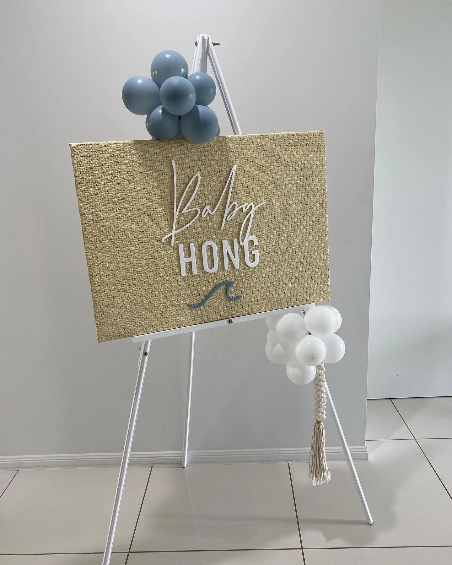 A decorative baby shower sign with a beige textured background, white and blue balloons, and the words 'Baby HONG' in white and black lettering, with a small wave graphic, standing on a white easel.