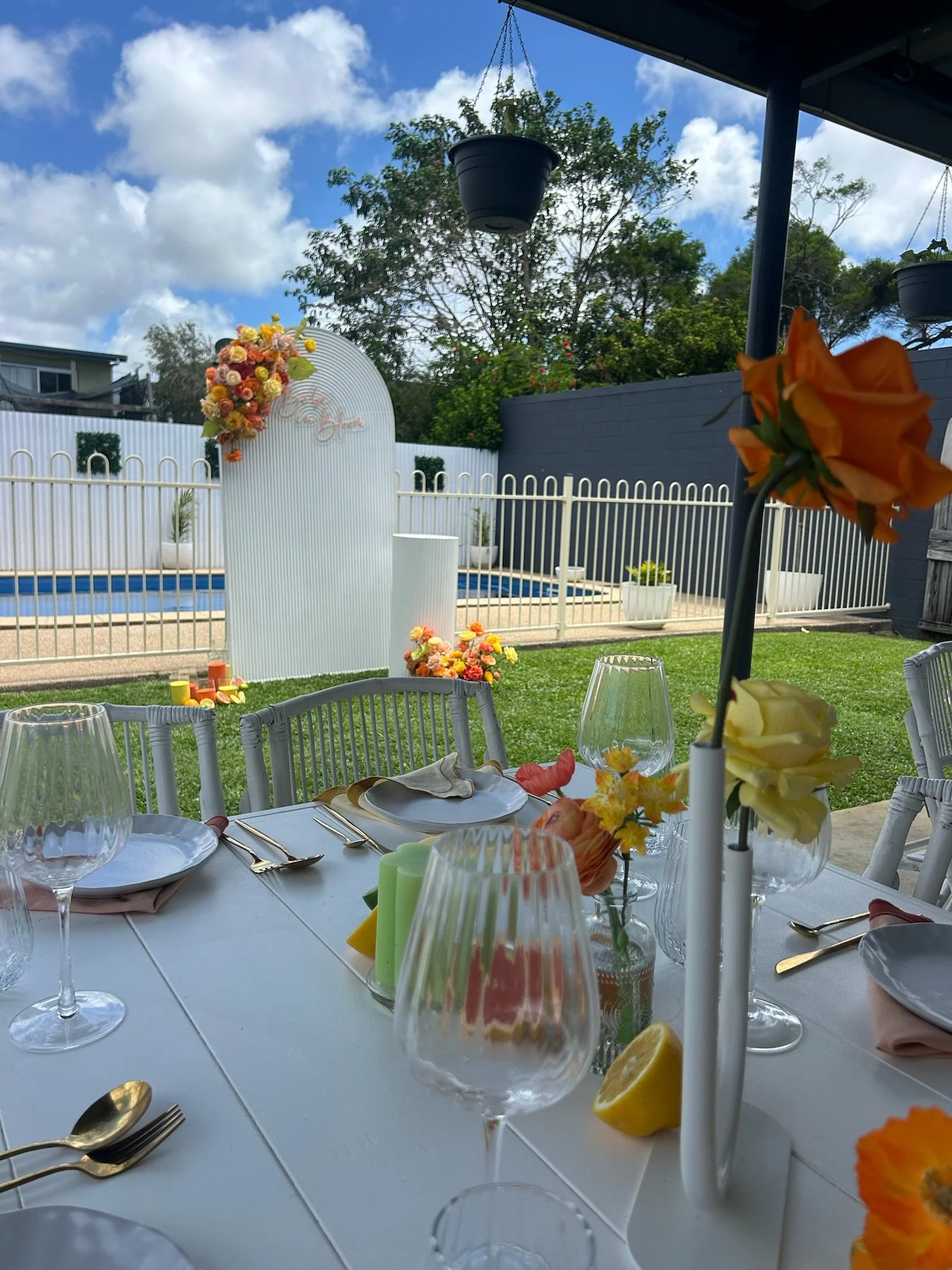 A decorated outdoor dining table with floral centerpieces and glassware, set near a pool with a white fence and a backdrop of trees and a partly cloudy sky.