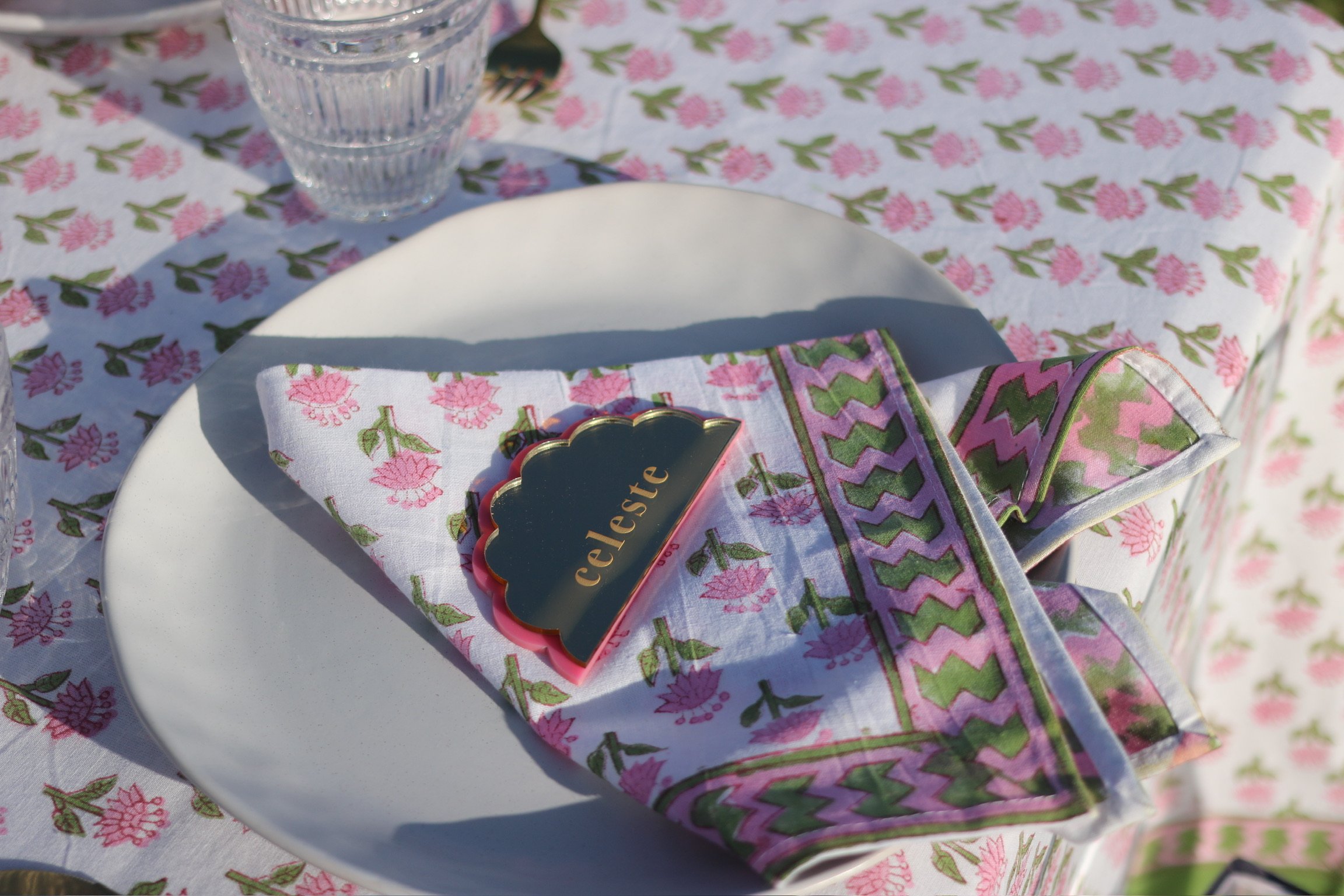 A table setting with a white plate, a folded cloth napkin with pink and green floral and zigzag patterns, a small black and gold badge reading "celeste," and a clear glass of water on a pink floral tablecloth.