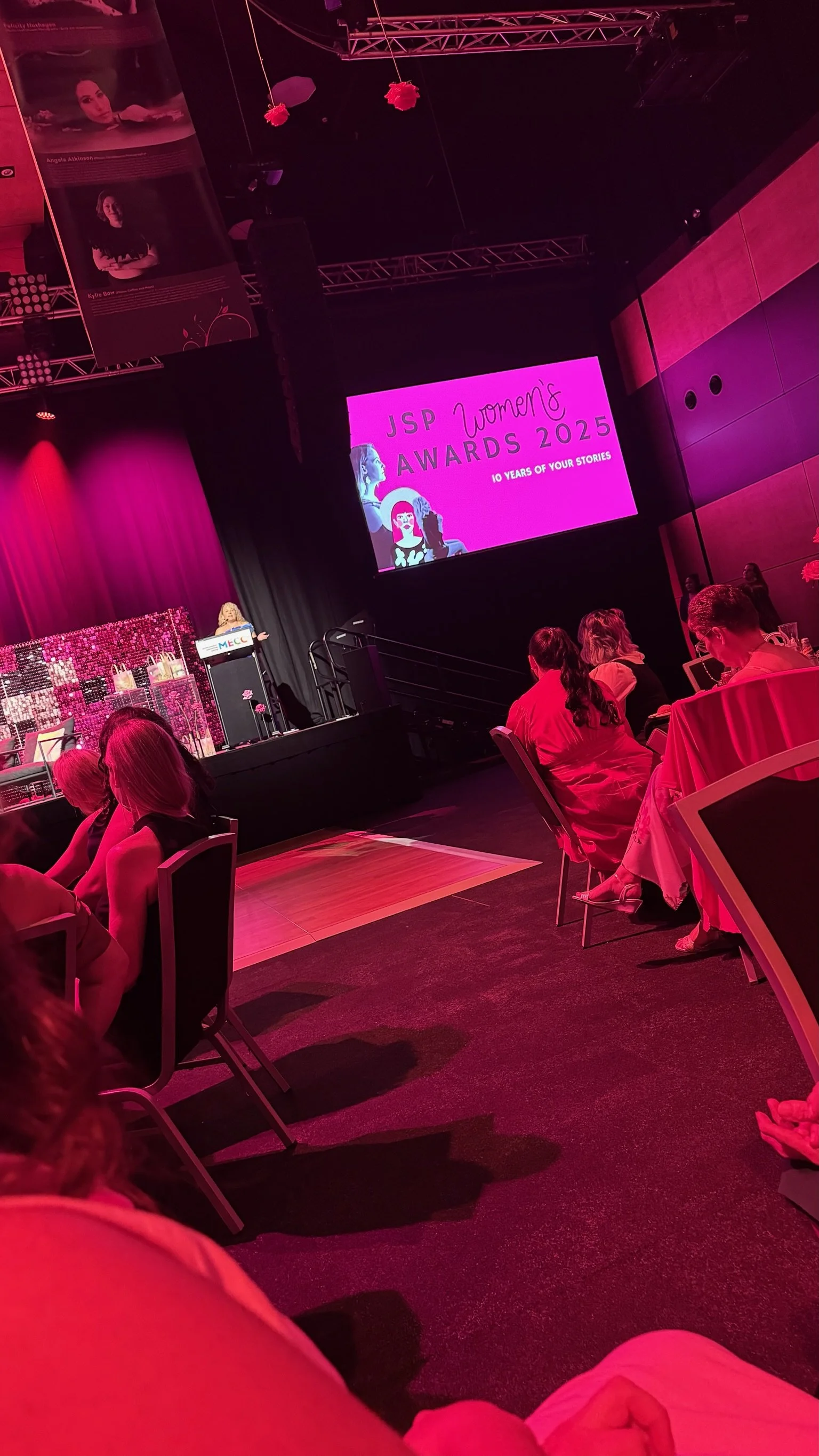 An indoor stage setting at the JSP Women's Awards 2023 event with a large pink screen displaying event details, a speaker at the podium, and women seated at tables, all illuminated by pink and purple lighting.