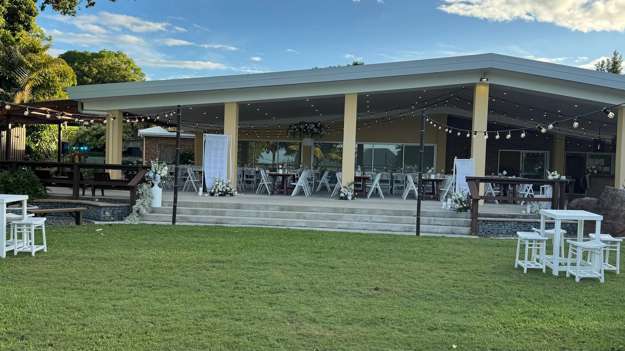 Outdoor event space with green grass, stairs leading to a covered patio with tables and chairs, string lights overhead, and decorative flowers and greenery.
